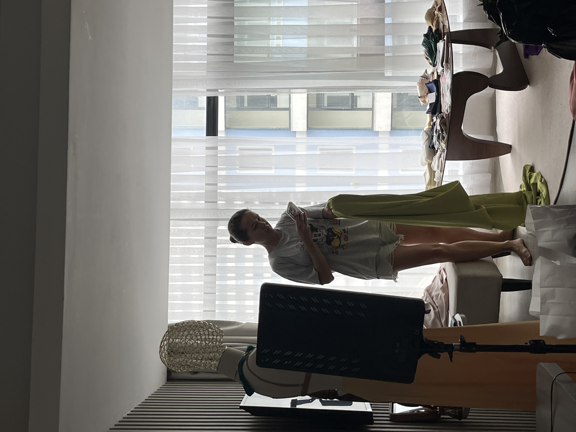 A woman is lying on a white surface, possibly a massage table or a similar cushioned surface, and using her cell phone. She is wearing a gray t-shirt and shorts. A green cloth drapes over her legs. There is a light source, possibly for photography or video recording, positioned in front of her. Behind her, a large window with sheer white curtains reveals a cityscape with buildings that appear to be located in an urban environment. Based on the mention of Bac Nha Trang Ward, Vietnam, the scene is presumably taking place in a building in that area. A round wooden table with miscellaneous items on top stands behind the woman’s head. There are also some items of clothing and other objects on the floor.