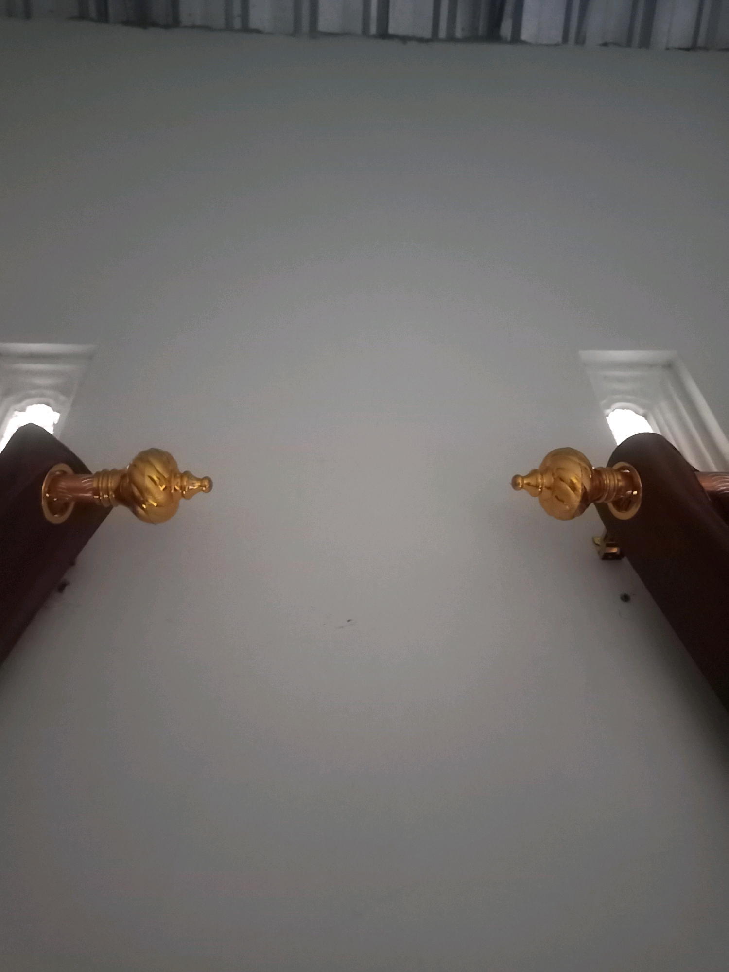 Two ornate golden finials are visible, attached to dark wooden fixtures. The finials have a segmented, bulbous shape with intricate detailing, suggesting a decorative purpose. They are positioned against a plain, light-colored wall. In the background, a ceiling with horizontal panels is visible, along with hints of what appear to be windows or light sources emitting a bright, diffused light. The scene has a static, architectural quality. There is no indication of people, animals, or actions occurring. The location is described as Karangtalun, Indonesia, but there are no specific visual cues in the image to confirm this.