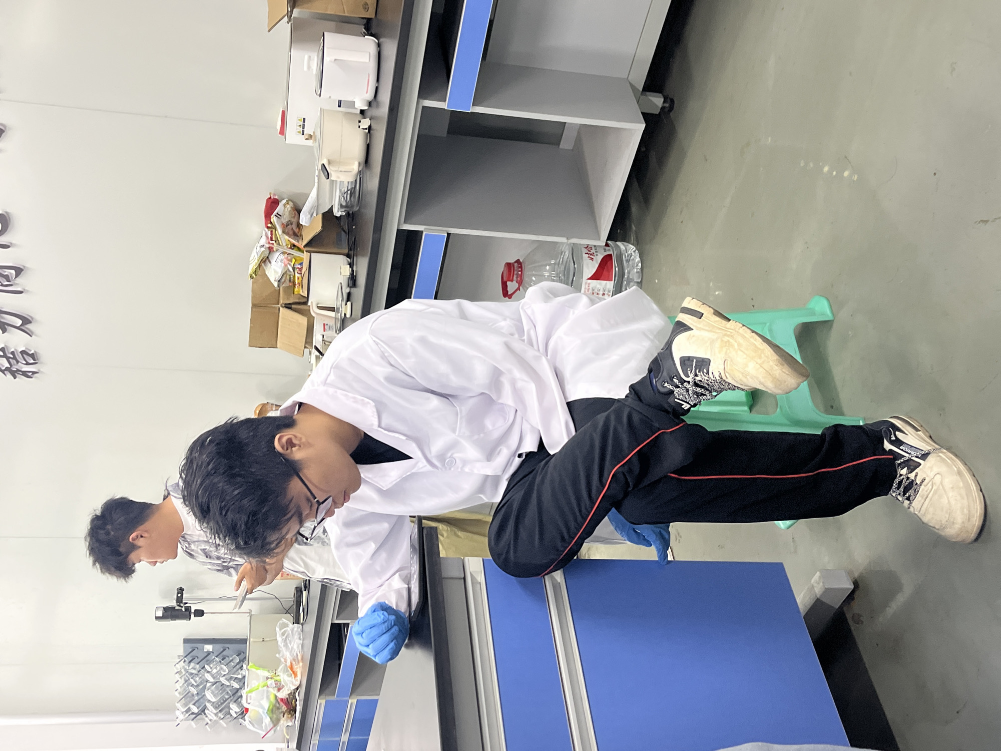 Two individuals, presumably male, are observed in what appears to be a laboratory setting. Both are wearing lab coats; one is also wearing glasses and blue gloves. The person wearing glasses is seen leaning over a blue countertop, while the other is standing behind, seemingly engaged in a task. Lab equipment and containers are visible in the background, along with shelves holding various items. The setting is located in Jiangjin, China.