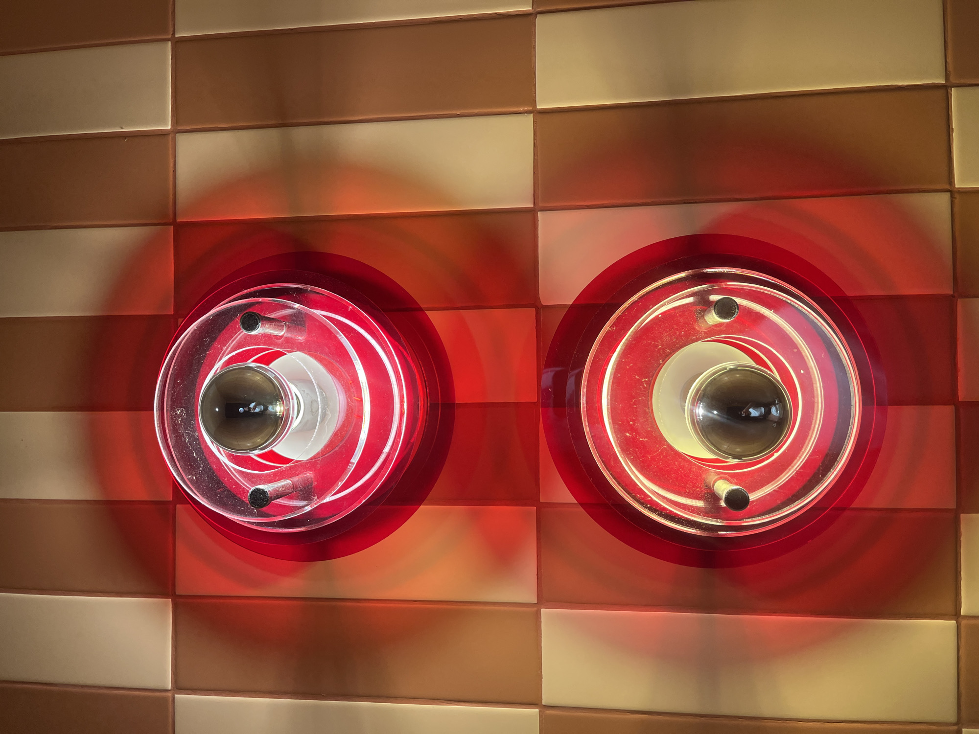 The image shows two illuminated, circular red light fixtures mounted on a wall of alternating light brown and dark brown rectangular tiles. The light fixtures have transparent, concentric rings surrounding a central bulb. The location appears to be Bac Nha Trang Ward, Vietnam, based on the prompt, although there are no visual cues within the image to confirm this. No people or interactions are visible.