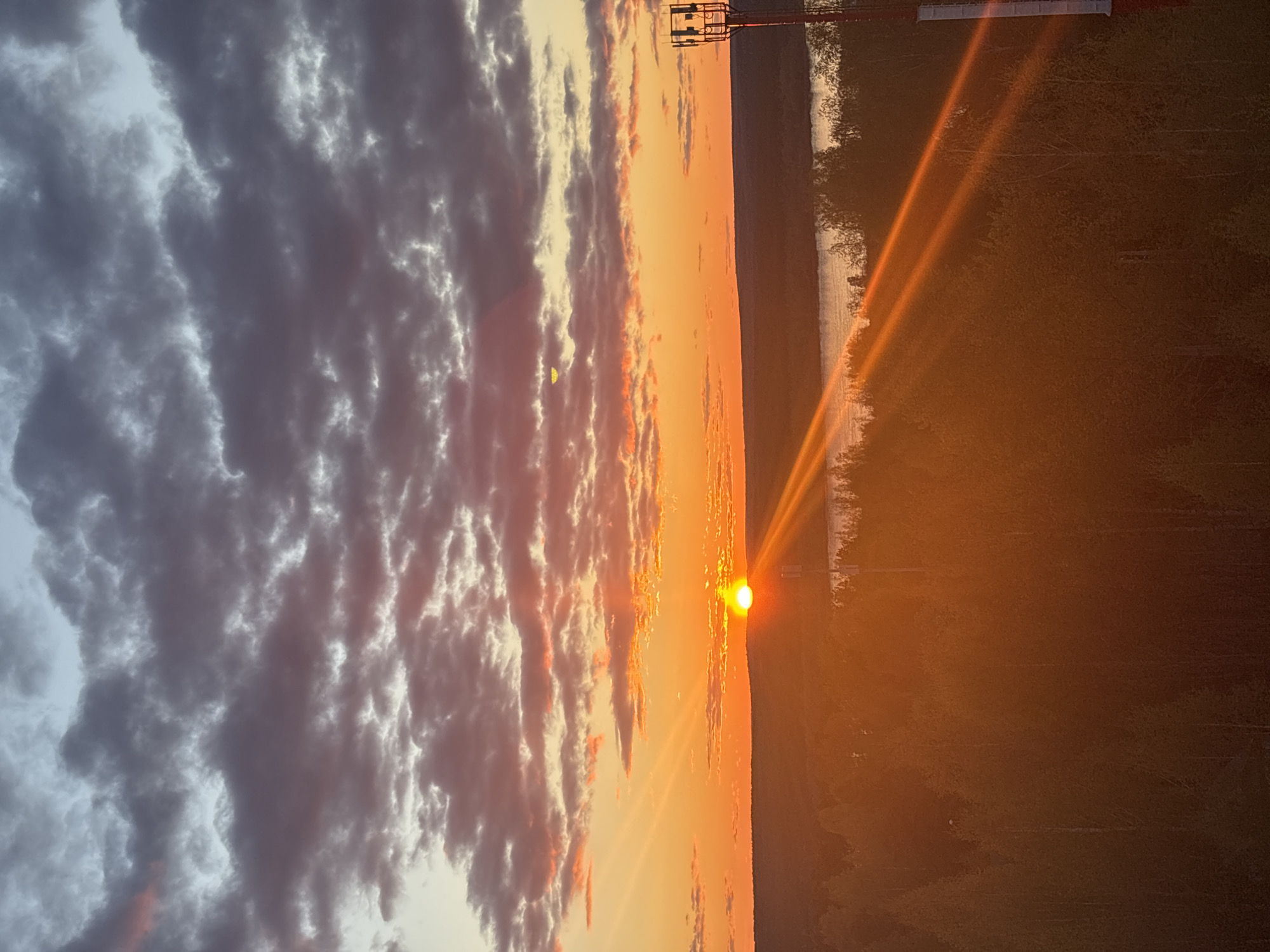 The image depicts a sunset over the Kama River in Perm, Russia. The sun is positioned on the horizon, emitting orange and red rays across the sky. Clouds are present on the left side of the image, ranging in color from grey to pink. A communication tower is visible on the right side of the image, situated on the bank of the river. The river itself is reflective, mirroring the colors of the sky. The surrounding area appears to be a wooded area with trees lining the riverbank.