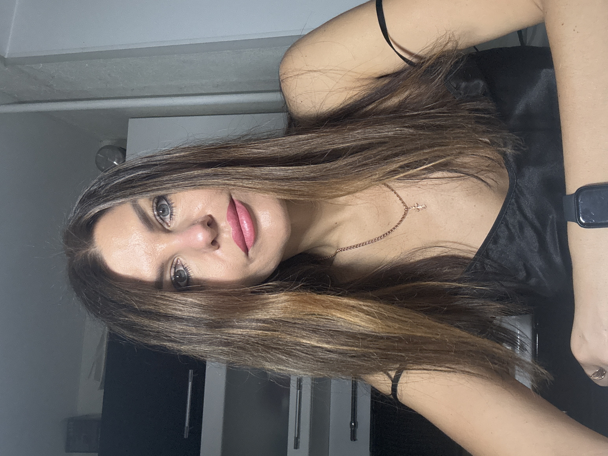 A woman with long brown hair and fair skin is pictured indoors. She is wearing a black top with thin straps and a necklace with a cross pendant. On her left wrist, she is wearing a smartwatch. The background includes a gray wall and a cabinet with black doors and silver handles. The location is indeterminate, and there is no information given about Chaikovsky, Russia.