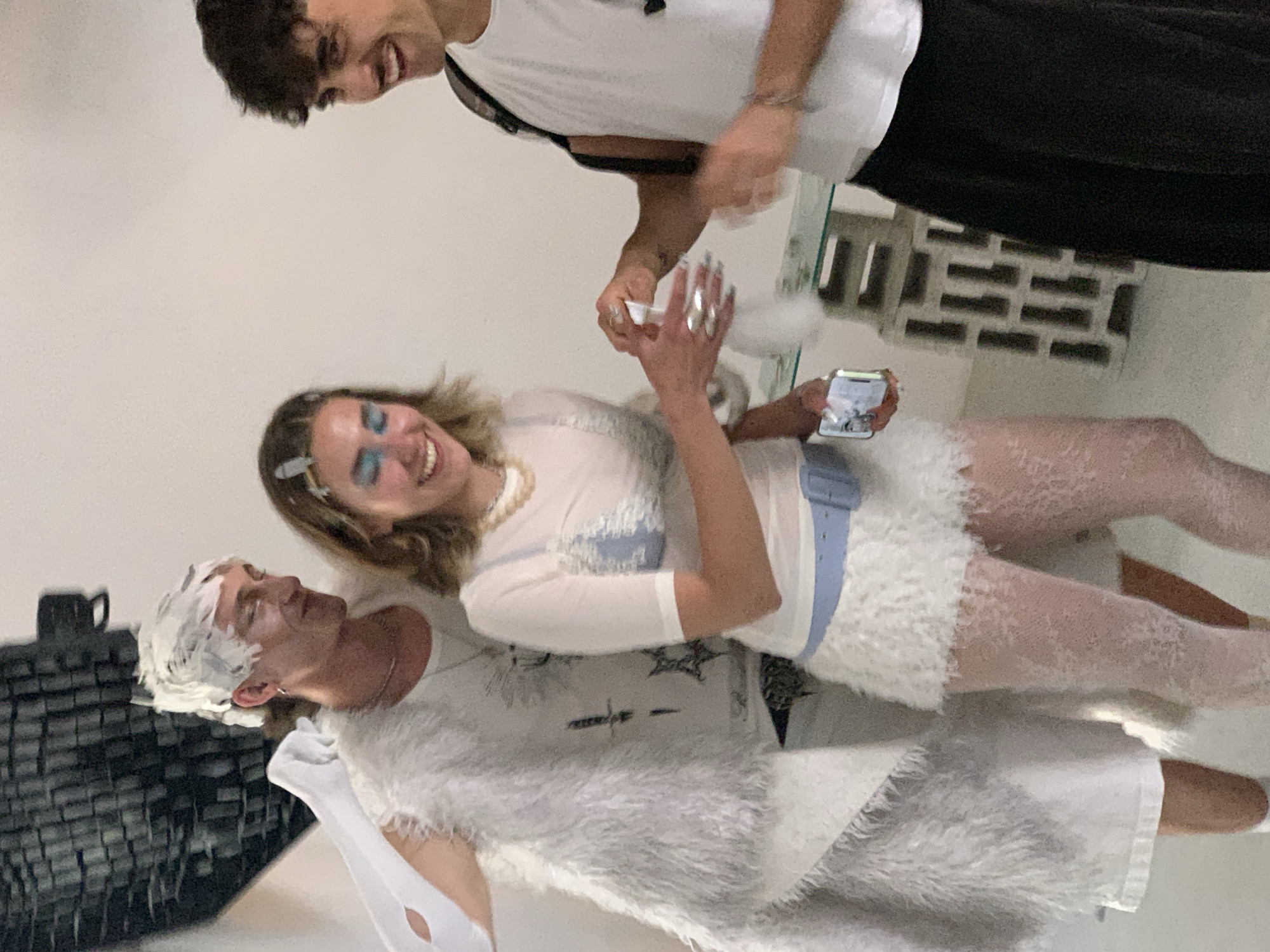 An indoor scene in Tel-Aviv, Israel, captures a lively and playful moment among three young individuals. The image, taken from a slightly elevated perspective, shows them engaged in a lighthearted interaction.
In the foreground, a young man is seen lying or leaning back, his head and short hair completely covered in white foam or cream. He wears a white t-shirt with a dark graphic design of stars and symbols, and a voluminous white, fluffy, faux-fur shawl or coat. His eyes are squinted shut or closed, and he has a smile on his face, indicating amusement.
Above and slightly behind him, a young woman with medium-length blonde hair, accentuated with a hair clip and prominent blue eyeshadow, actively applies more of the white foamy substance. She wears a white long-sleeved top with light blue details, a white fuzzy mini-skirt, and white lace stockings. A light blue belt, a pearl necklace, and several large silver rings on her fingers complete her distinct outfit. She holds a white smartphone in her left hand, displaying a wide, joyful smile.
Further back and appearing to look down into the scene, another young man with short dark curly hair and a white t-shirt smiles broadly. All three subjects exhibit expressions of joy and mirth, suggesting a fun, jovial atmosphere, possibly during a party or a game.
The background consists of plain white walls. Decorative elements include a dark, slatted or brick-like structure on the left and a lighter, patterned brick-like divider on the right. The lighting appears to be artificial indoor illumination, and there is no visible text.