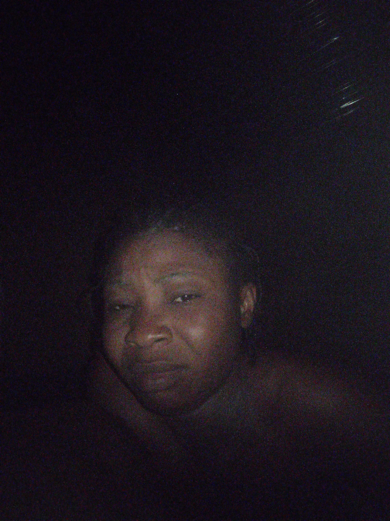 A dark, grainy image shows a person with brown skin. The person has their hair pulled back, and the face appears to have a somber expression. The environment is undefined and very dark. The location and other environmental details are not discernible.