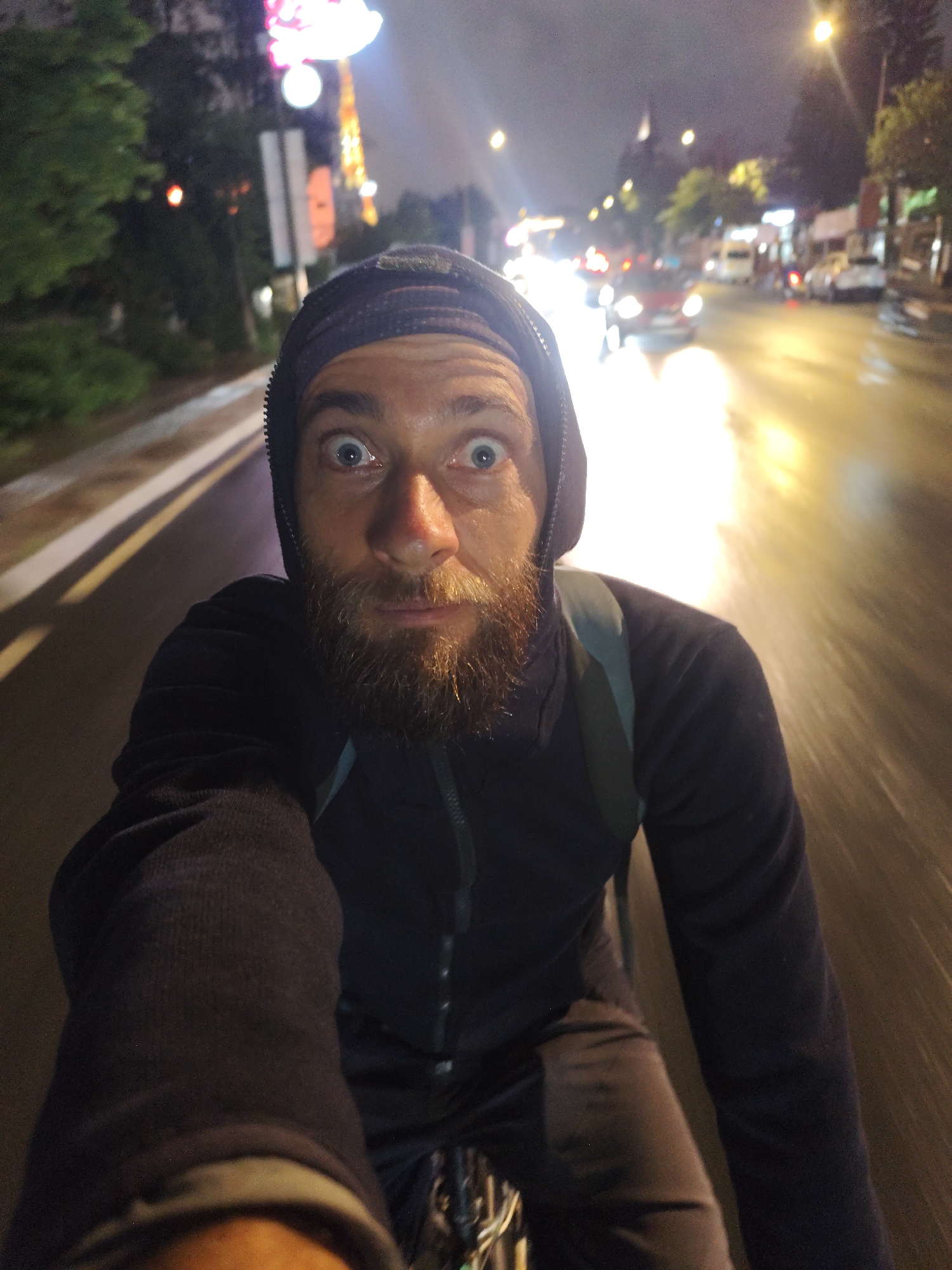 A man with a beard is riding a bicycle at night in Da Lat, Vietnam. He is wearing a dark hooded sweatshirt and a backpack. The road is wet and reflects the lights of the city. There are cars and buildings visible in the background. The man is looking directly at the camera with a surprised expression. The image is slightly blurred, suggesting motion.