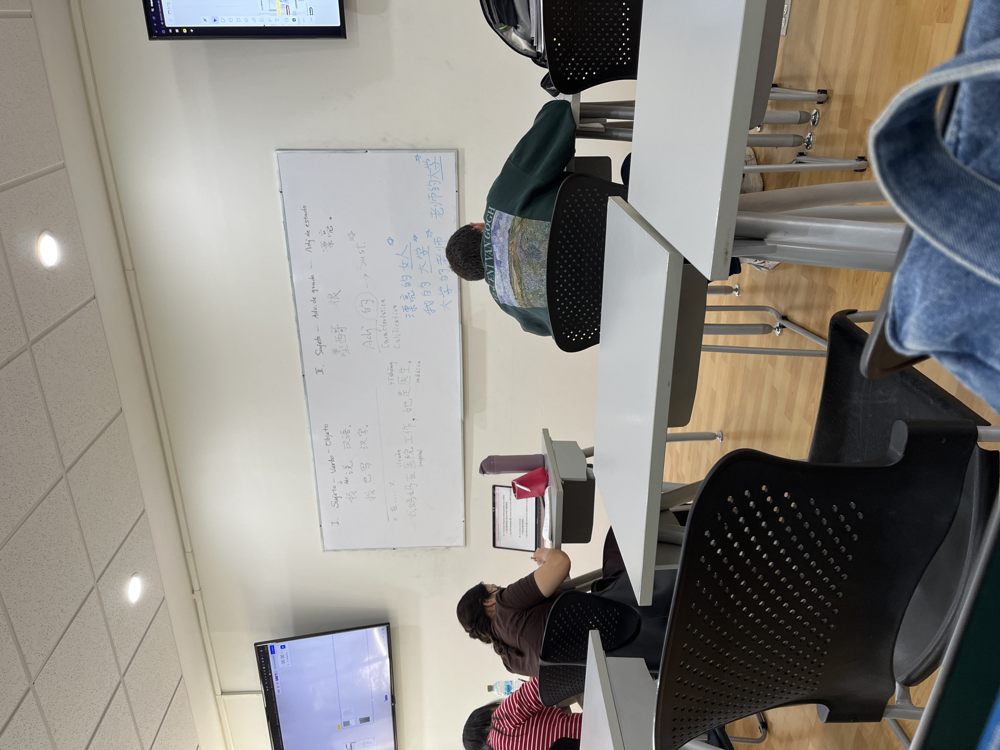 The image shows a classroom setting with students seated at desks. A whiteboard displays notes written in both English and Mandarin Chinese, covering grammar concepts and vocabulary related to adjectives, subjects, verbs, and objects. One student is using a tablet device. There are two monitors, one displaying a Miro board, in the room. The location of the image is a classroom, likely in a language learning center, in San Bernardino Tlaxcalancingo, Mexico.