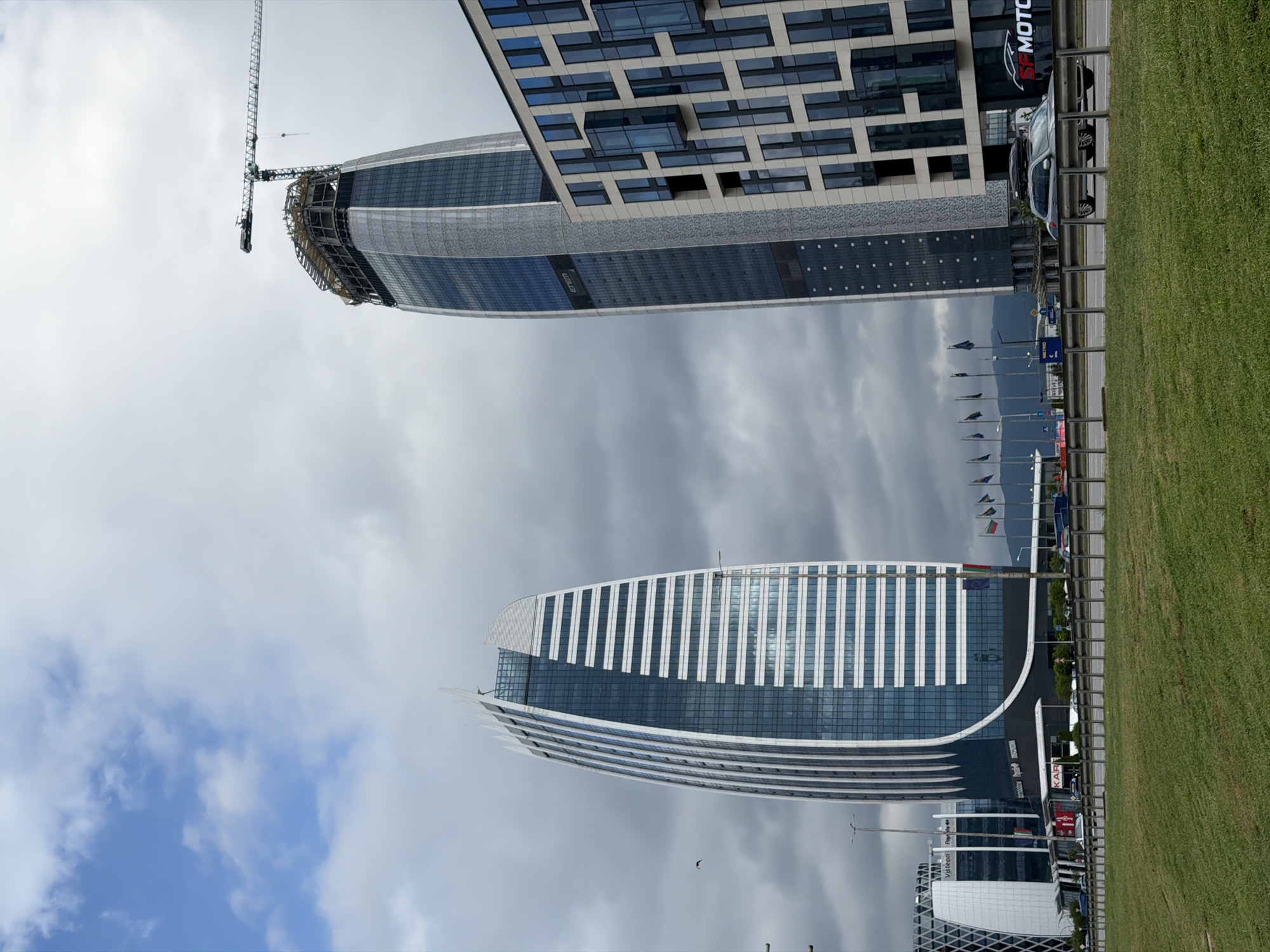 The image captures two modern, curved buildings in Sofia, Bulgaria. The lower building is mostly complete, featuring horizontal white stripes and blue-tinted glass. The upper building is under construction, with a crane visible at the top. The sky is cloudy. The buildings are separated by a road, with flags and parked cars alongside a grass area. A car dealership with the name "MOTOR" is visible on one side.