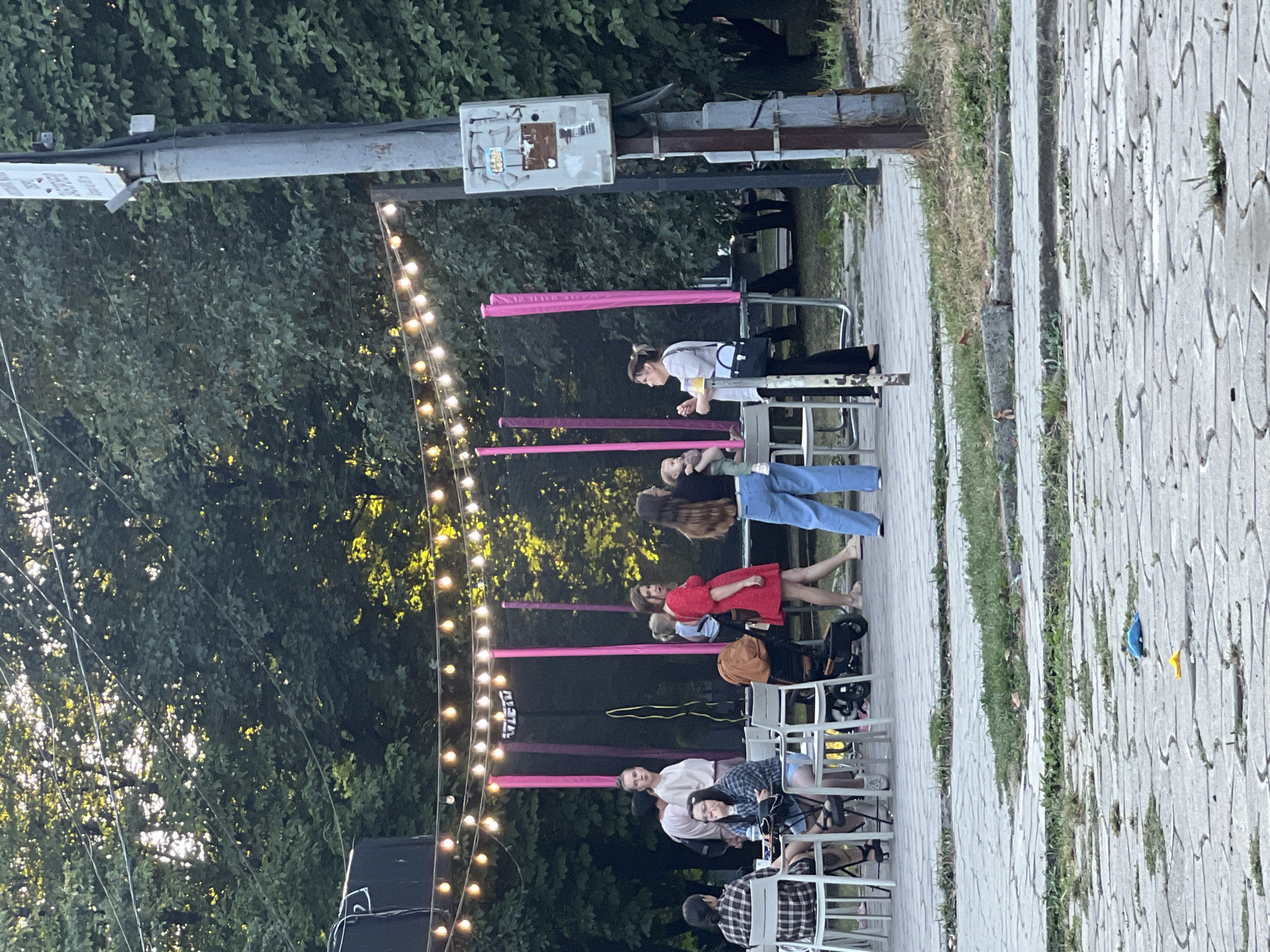 This image depicts an outdoor gathering at dusk, possibly a cafe or a public space in Sheptytskyi, Ukraine. A string of lights hangs overhead, illuminating a group of people. In the foreground, a paved area with some scattered debris leads to a grassy embankment.

Several individuals are visible. Two people are seated at a table on the left, appearing to be engaged in conversation. A woman in a red dress stands near the center, while another woman holds a baby. A stroller is positioned behind them. Further back, a large trampoline with pink supports is visible. A utility pole with signage stands to the right of the frame. The background is dominated by lush green trees, suggesting a park-like setting or a street lined with foliage. The overall lighting indicates late afternoon or early evening.
