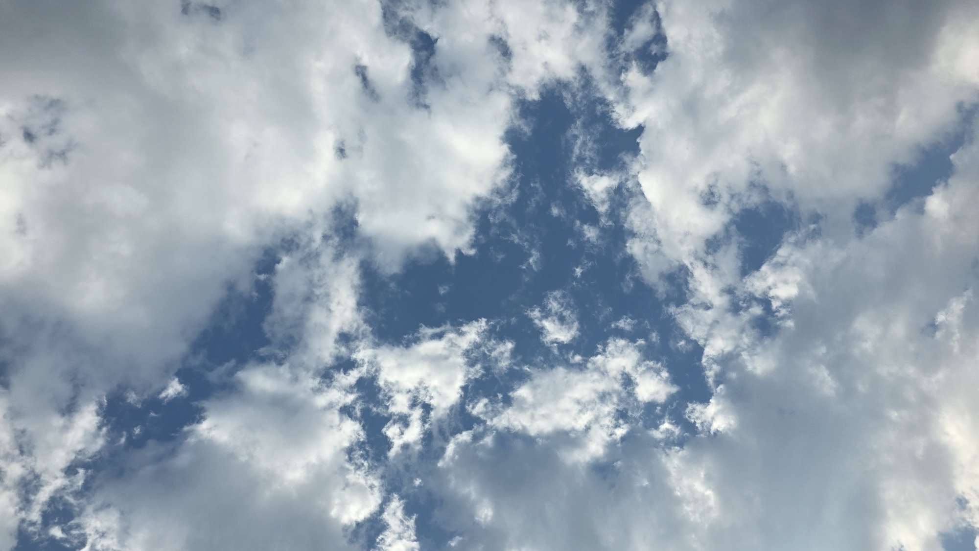 The image is a vertical shot of a blue sky filled with scattered white and gray clouds. The clouds vary in density, with some appearing thin and wispy, while others are more substantial and opaque. The overall impression is that of a partly cloudy day. The photograph does not depict any elements of the city of Chuy, Kyrgyzstan; it is solely an image of the sky.