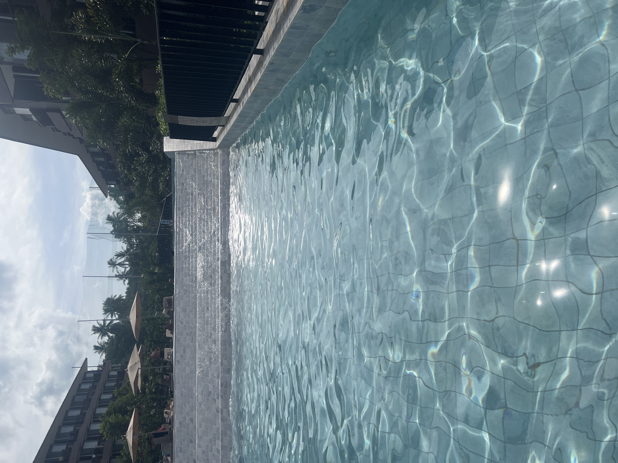 A wide shot captures a swimming pool in Ko Samui District, Thailand, with tiered steps leading up to a resort building. The pool water is clear blue with sunlight reflecting off its surface, revealing the tiled bottom. Along the edge of the pool, a dark metal fence runs parallel to the resort's facade. Several beige umbrellas are positioned on the upper tier of the steps, providing shade for lounging guests. The resort building, constructed with dark wood and windows, is surrounded by lush green trees and palm trees, indicating a tropical setting. The sky above is partly cloudy, with a hint of blue peeking through.