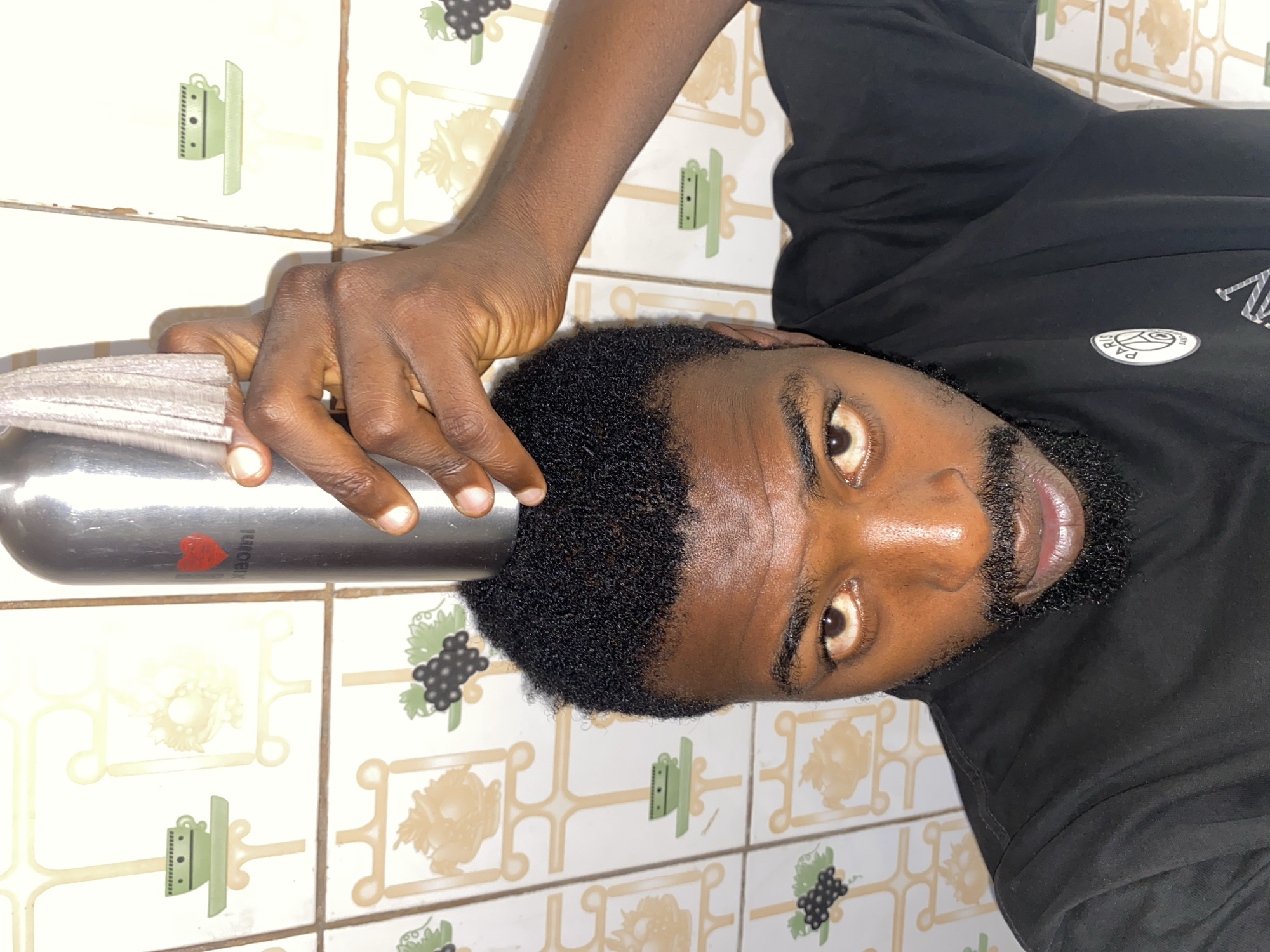 A medium-shot, close-up photograph shows a Black man with dark hair and a beard, wearing a black long-sleeved shirt with a circular logo on the chest. He is holding a metallic water bottle in his left hand, with the open end covered by a gray cloth. The background consists of patterned tiles with images of kitchenware and grape bunches. There is no evidence in the image to verify the location as Jos, Nigeria.