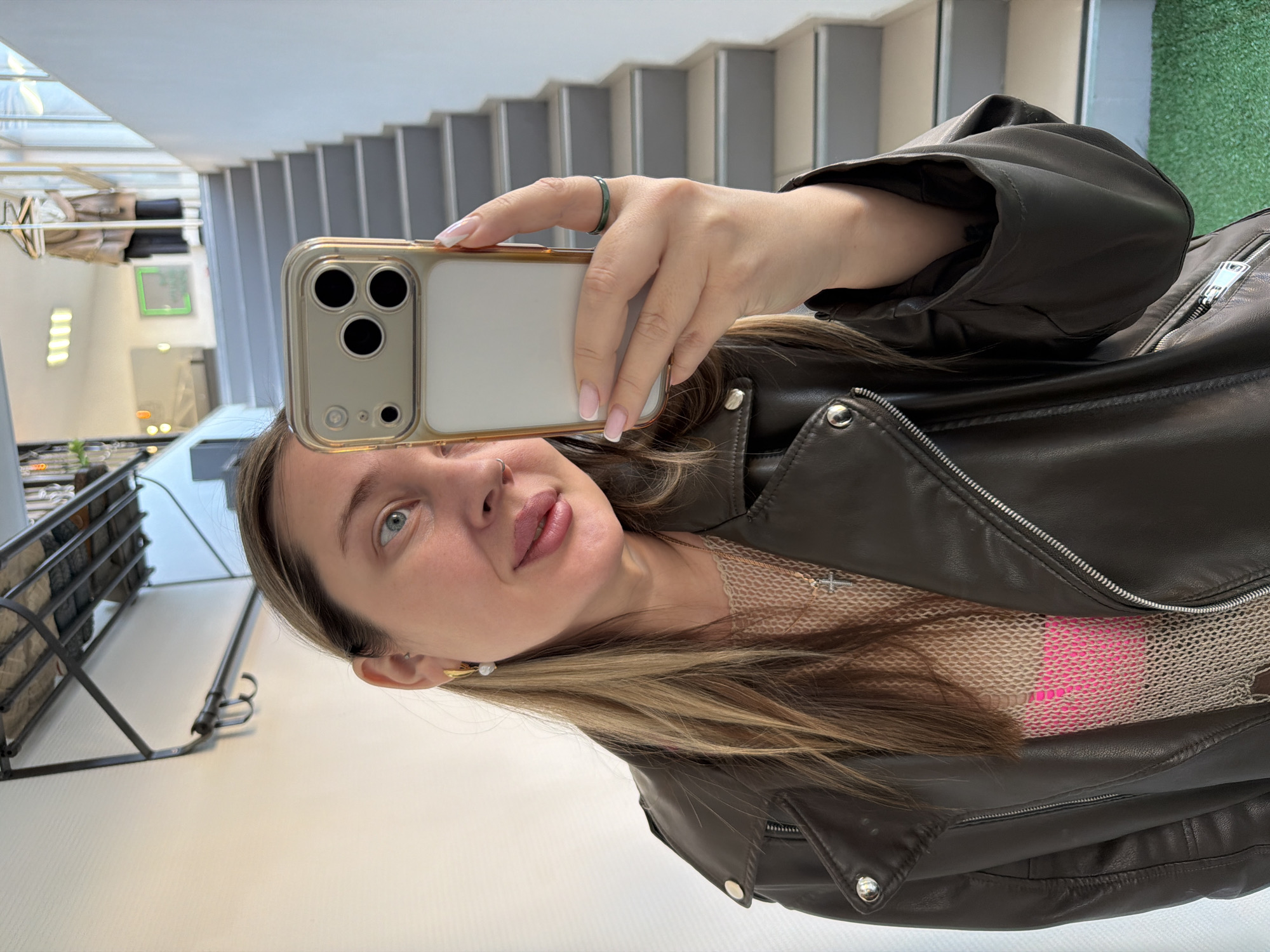 A woman wearing a brown leather jacket is taking a selfie with a gold-cased phone. The image is taken indoors near a set of stairs. The reflection on the phone case shows a clothing rack and possibly part of a store. The location is likely in Chaikovsky, Russia.