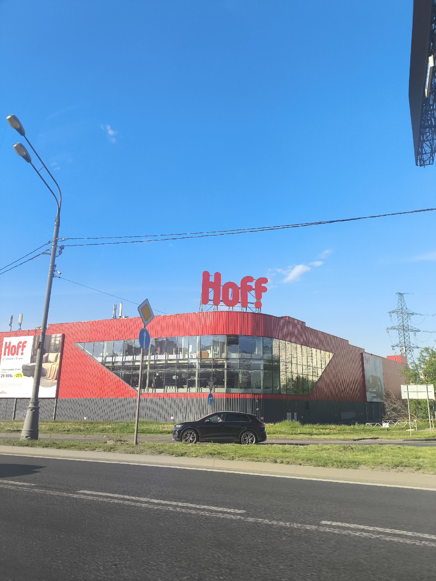 The image shows a Hoff store on a clear day in an unknown city. The building is red and grey with large glass windows. The Hoff logo is prominently displayed on the roof. A black SUV is driving on the road in front of the store. A street light stands to the left. Overhead power lines are visible against the clear blue sky. A power tower is visible in the background.
