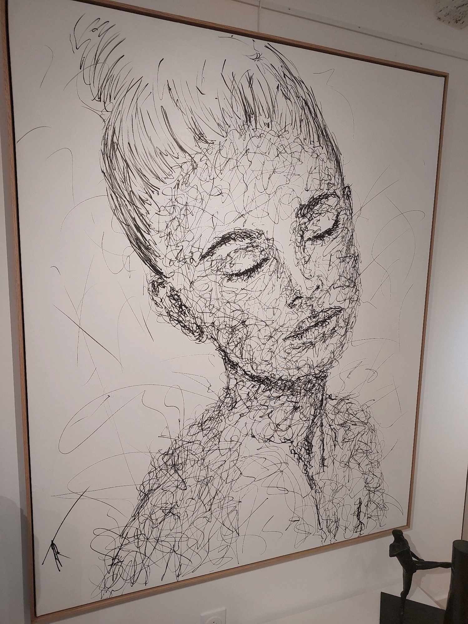 The image depicts a large, framed ink drawing of a woman's face rendered in a chaotic, scribbled style. The drawing is in black ink on a white background, with the lines forming a dense, almost abstract representation of the subject's features, including closed eyes, nose, lips, and hair pulled up. The frame is simple, light-colored wood. The artwork is hanging on a white wall, possibly in an art gallery or studio. In the lower right corner, a small, dark bronze sculpture of a figure in a dynamic pose is visible on a dark pedestal. The lighting suggests an indoor environment. This artwork is on display at the Fondation Maeght in Saint-Paul-de-Vence, France, a renowned museum dedicated to modern and contemporary art.