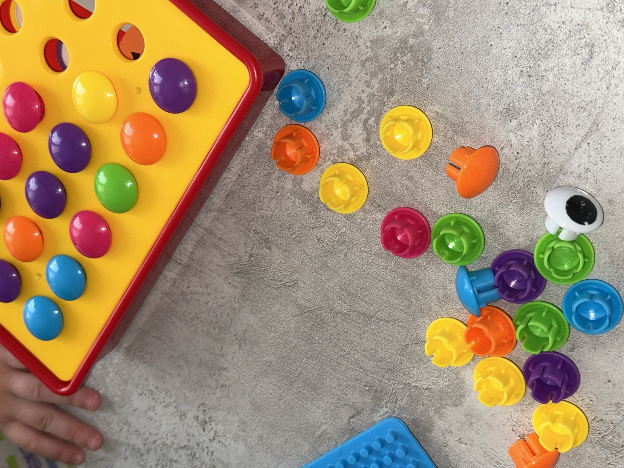 The image shows a child's hand near a yellow and red pegboard with several colored pegs inserted. Loose colored pegs (blue, orange, yellow, pink, green, and purple) are scattered across a grey textured surface, along with a blue rectangular object with circular indentations. One peg has a cartoon eye on it. The location in the city of Chaikovsky, Russia is unknown.