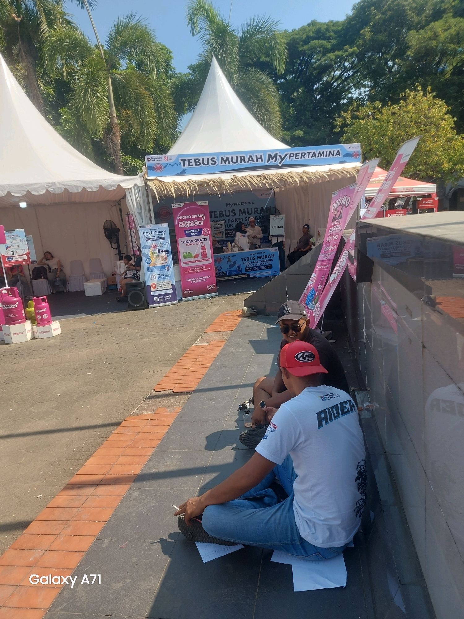 This is an outdoor daytime photograph taken at an event featuring tents, likely a promotional or sales event. The scene is set in Makassar, Indonesia.
In the foreground, two men are sitting on the ground next to a low, gray wall. The man on the left is wearing a dark baseball cap, sunglasses, and a dark t-shirt. The man on the right is wearing a red cap with "Arai" visible on it, and a white t-shirt with the word "RIDER" printed in large blue letters across the back. Both men are looking towards the left. Papers are spread out on the ground in front of them. The man on the right has a cigarette in his right hand.
Behind the seated men, there are several white tents, some with thatched roofs. Banners and posters are visible, advertising "MY PERTAMINA" and "Bright Gas." One banner states "TEBUS MURAH MY PERTAMINA". Pink gas cylinders are displayed in front of one of the tents. Inside the tents, people can be seen sitting and standing, and there is a black speaker.
In the background, there are tall palm trees and other foliage, suggesting a tropical or subtropical environment. The sky is clear and blue. The ground in front of the tents is paved with gray tiles and bordered by orange brickwork.
The overall impression is a lively outdoor event, possibly a fuel or gas promotion, on a sunny day in Makassar.