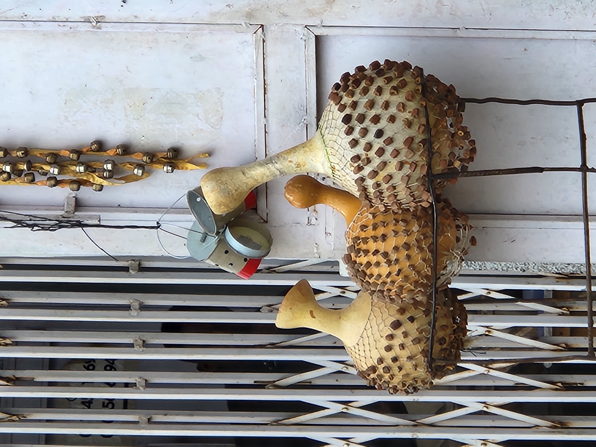 A horizontal, eye-level shot shows a closed storefront or entrance in Jos, Nigeria, featuring an unusual decorative arrangement. The primary feature is a set of three stacked, gourd-like objects covered in netting and studded with small, brown components. These objects appear handcrafted or artfully designed, possibly for sale or display. Adjacent to the gourds is a string of metal bells or small, spherical ornaments hanging from the ceiling. Further left, a cylindrical object with metal components and a red accent is attached to the ceiling. The storefront has a rolling, slatted metal gate and a white, paneled wall behind the arrangement. The overall aesthetic suggests a local artisan's shop or a decorative display specific to the culture and location.