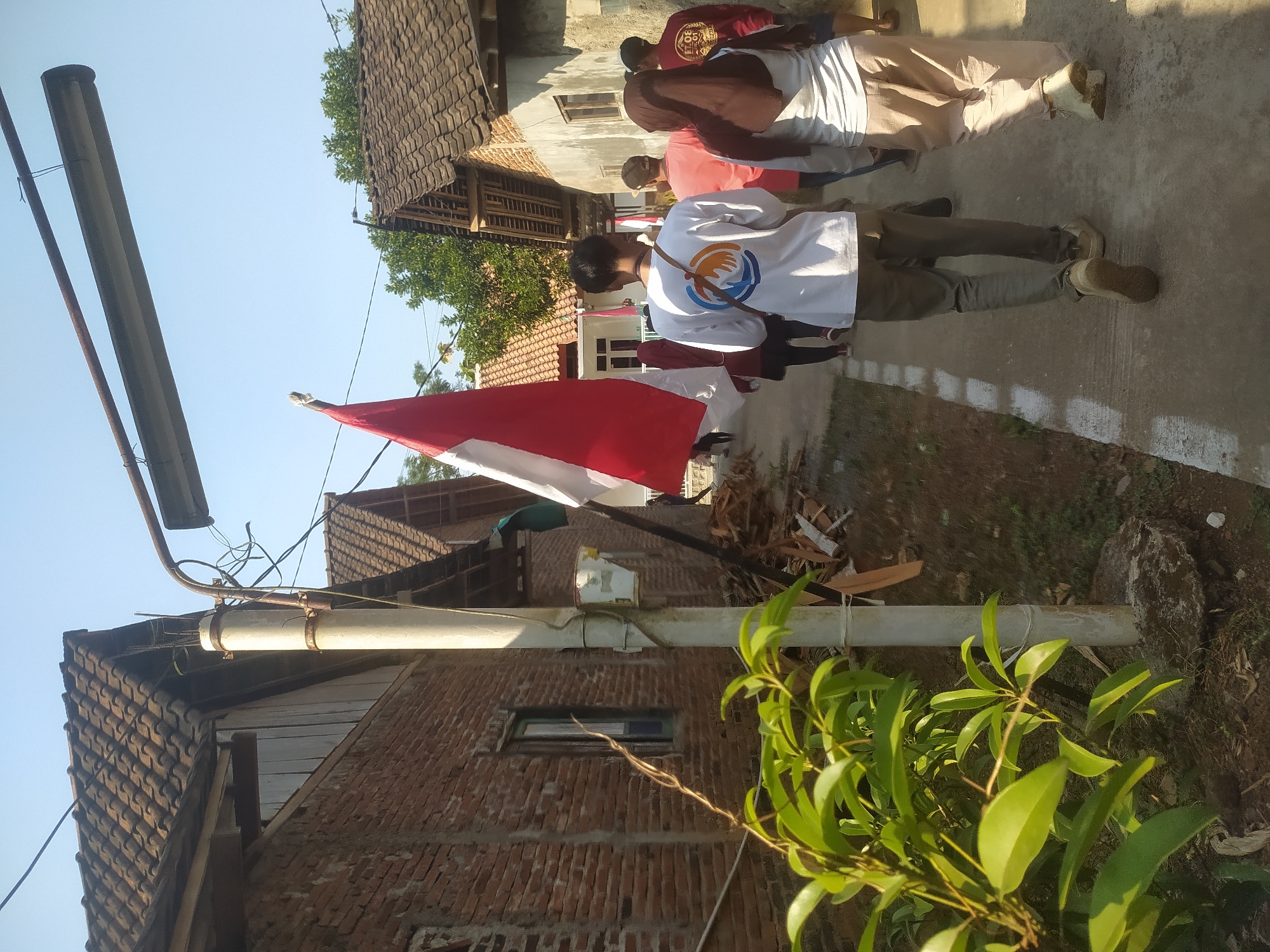 A group of people, some carrying an Indonesian flag, walk along a narrow path in a rural village. The path is bordered by brick buildings with tiled roofs on either side. The flag is red and white, and it is being held aloft by a person in the foreground. The people are dressed in casual clothing. The sky is clear and blue. The scene appears to be set in Tuntang, Indonesia. The overall impression is one of a community event or procession.