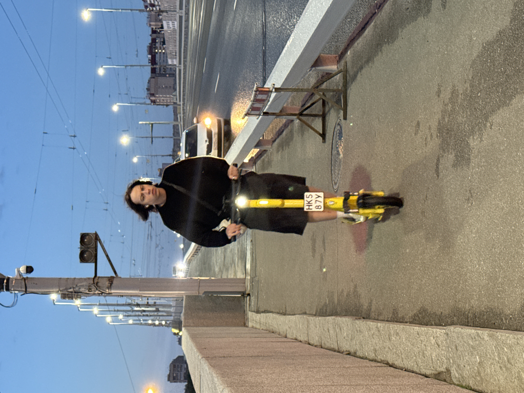 The image depicts a street scene in Kudrovo, Russia, possibly on a bridge, during dusk or early evening. A young person with shoulder-length brown hair, wearing a black long-sleeved shirt and dark shorts, is riding a yellow electric scooter. The scooter has a license plate that reads "HK5 87Y". The person is wearing white socks and white sneakers. The scooter has a bright headlight illuminated.
The background features a road with a white car approaching, its headlights on, and other vehicles further in the distance. The sky is a deep blue with some clouds, and streetlights are visible along the roadside, casting a warm glow. On the left side of the image, there is a concrete wall with a textured surface. In the mid-ground, a white railing lines the road, with a temporary barrier featuring red and white elements placed on the pavement. The pavement appears wet in places, suggesting recent rain. The overall environment is urban, with buildings visible in the far distance.
