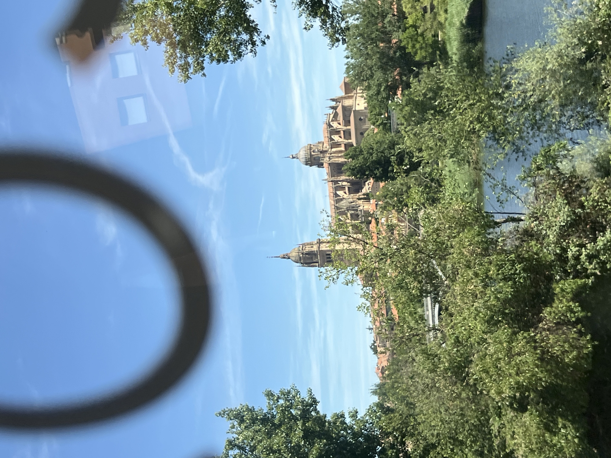 The image captures a view of the New Cathedral of Salamanca, Spain, seen from across the Tormes River. In the foreground, lush green trees and foliage dominate the scene, with a body of water reflecting the bright blue sky. The New Cathedral, a magnificent architectural landmark, stands prominently in the middle ground, its dome and spires reaching towards the sky. The building's stone facade is illuminated by the sunlight, highlighting its intricate details. The sky is a vibrant blue, dotted with a few wispy white clouds. There are no people visible in the image. The overall impression is a serene and picturesque landscape of a historic city.