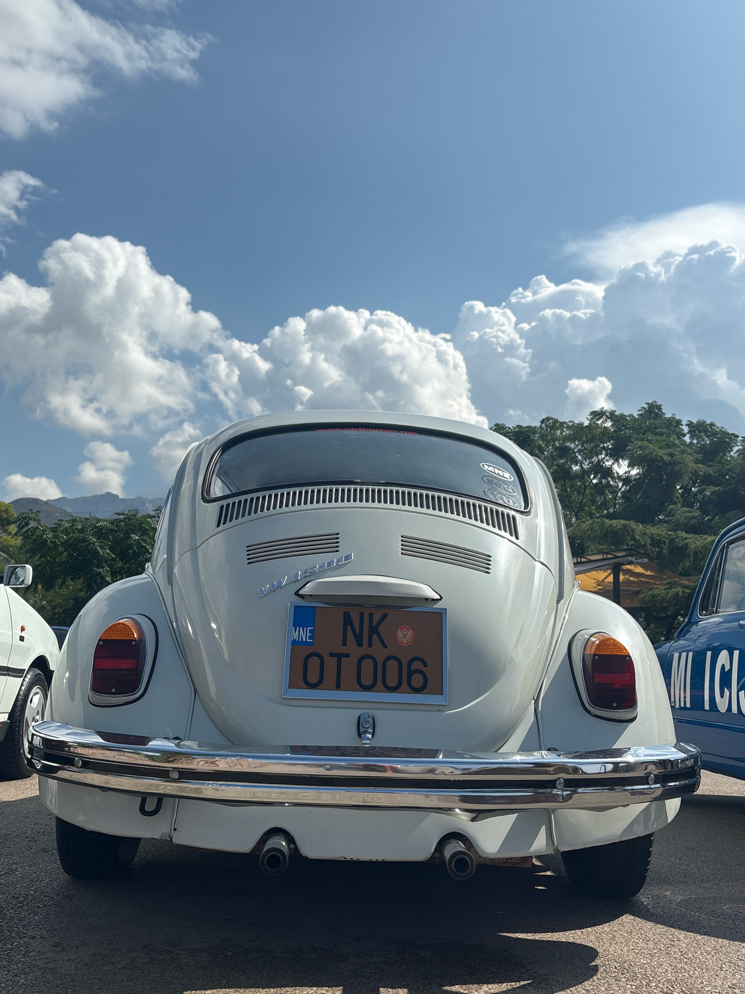 This is a rear view, eye-level shot of a white Volkswagen Beetle parked outdoors. The car has a chrome bumper and a license plate that reads "MNE NK OT 006". The license plate indicates the vehicle is registered in Montenegro. In the background, there are trees and a mountain range under a bright blue sky with scattered white clouds. To the right of the Beetle, a portion of a blue car with "MLICIC" visible on its side is present. To the left, the rear of a white car is partially visible. The scene appears to be a sunny day in the city of Bar, Montenegro, possibly at an outdoor car show or event. There are no people visible in the image.