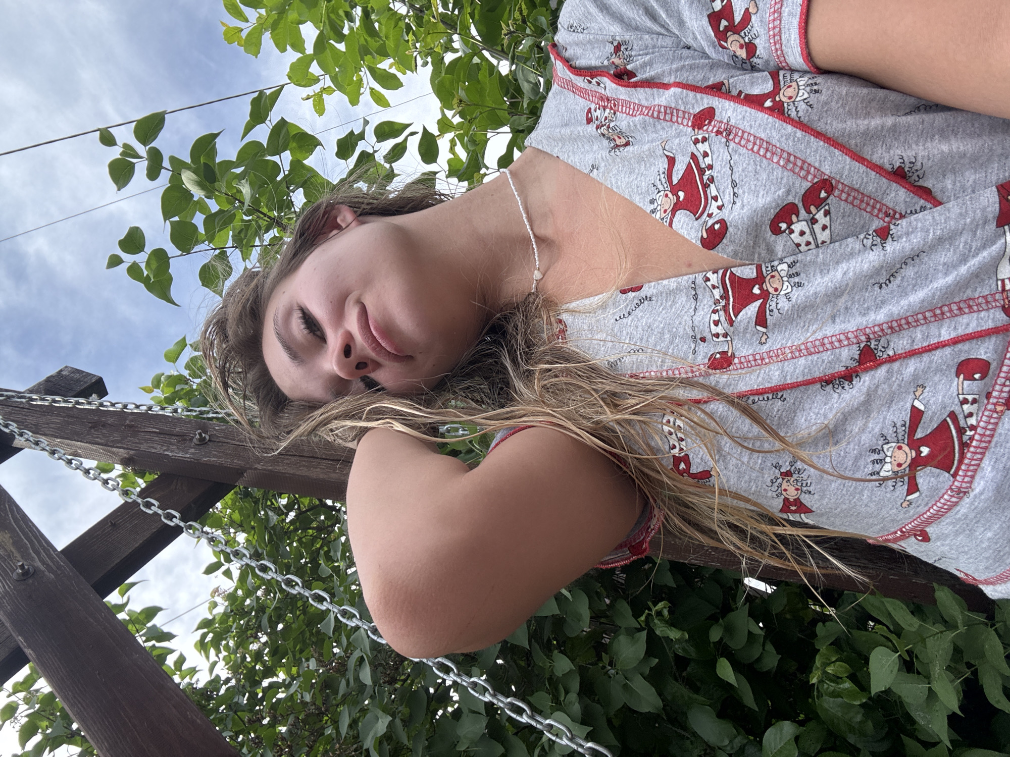A young woman lies on a wooden structure, possibly a swing set or a bench, in an outdoor setting. She is positioned in a way that her upper body and head are visible, while her lower body is out of frame. The location is likely outdoors, given the presence of lush green foliage, trees, and a glimpse of the sky. The woman is wearing a grey top with a repeating cartoonish pattern of figures in red dresses and white tights. She is also wearing a white beaded necklace. A chain and wooden beams indicate that she is either resting on a swing set or next to it. The location in Chaikovsky, Russia is not directly apparent from the visual information.