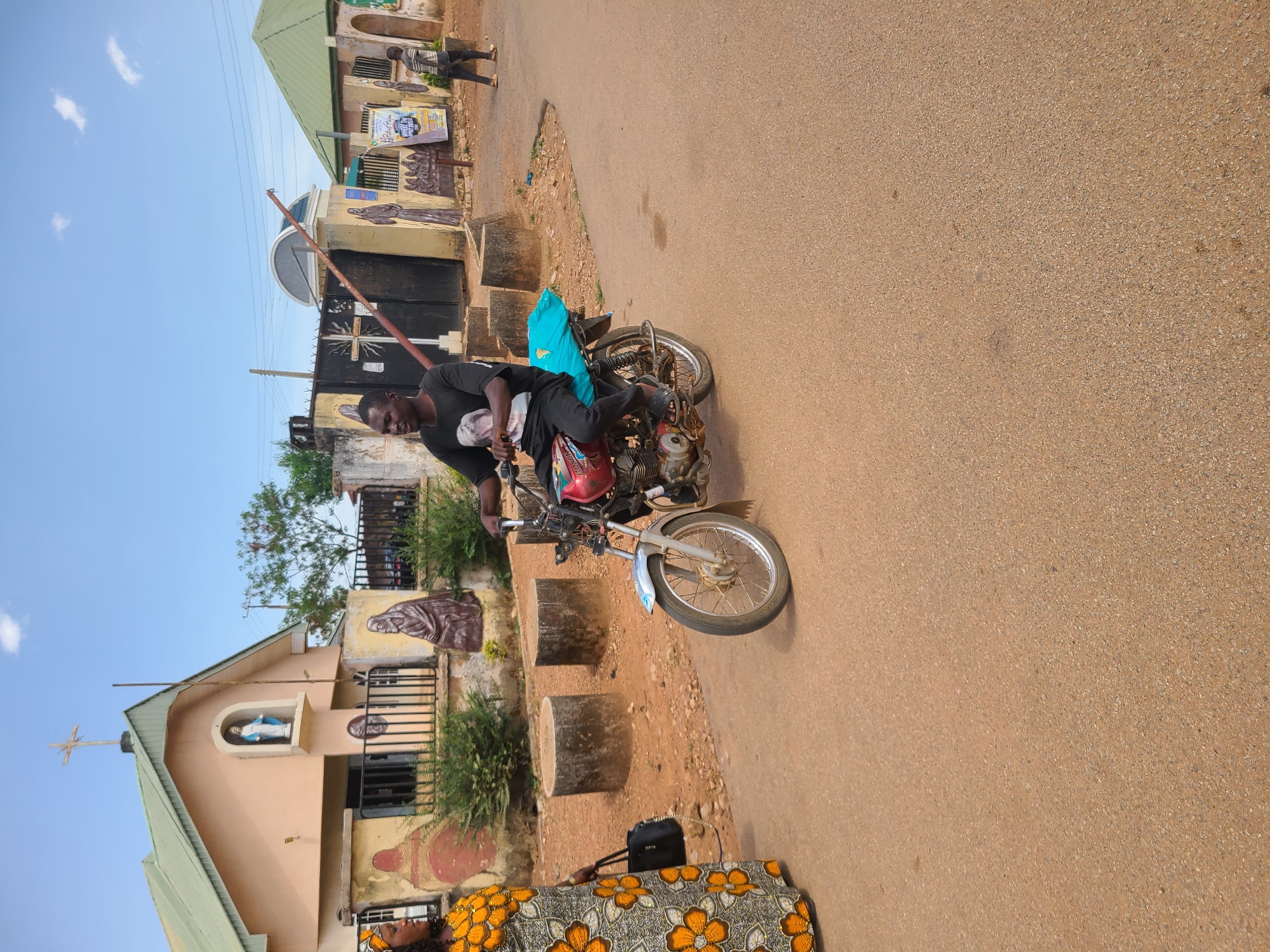 A man in a black t-shirt is sitting on a red motorcycle on a brown road. The background shows a church with religious iconography on the walls and a green roof. A person is walking in the distance. The scene is located in an urban area, with a gate and trees visible in the background. The city and location are unknown.
