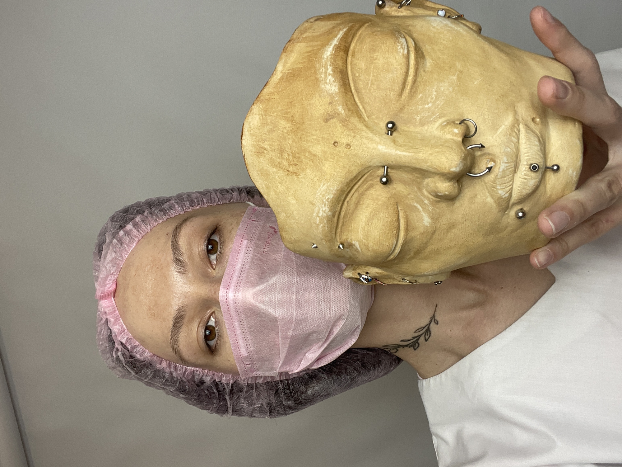 A person wearing a pink surgical mask and hairnet is holding a light-skinned face mask adorned with multiple piercings. The face mask is held to the side, partially obscuring the person's face. The person has brown eyes. A tattoo is visible on the person's neck. The background is plain and neutral. The image was taken in Izhevsk, Russia.