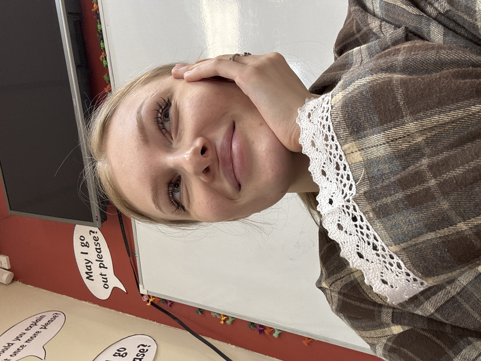 A blonde woman wearing a plaid shirt with lace trim on the collar poses for a selfie. Her face is tilted towards the camera, and she is smiling. Her left hand rests on her forehead. The background includes a whiteboard, a red wall, and speech bubble decorations. One speech bubble reads, "May I go out please?". The image location appears to be a classroom. There is no information available to determine the location of the image in the city of Perm, Russia.