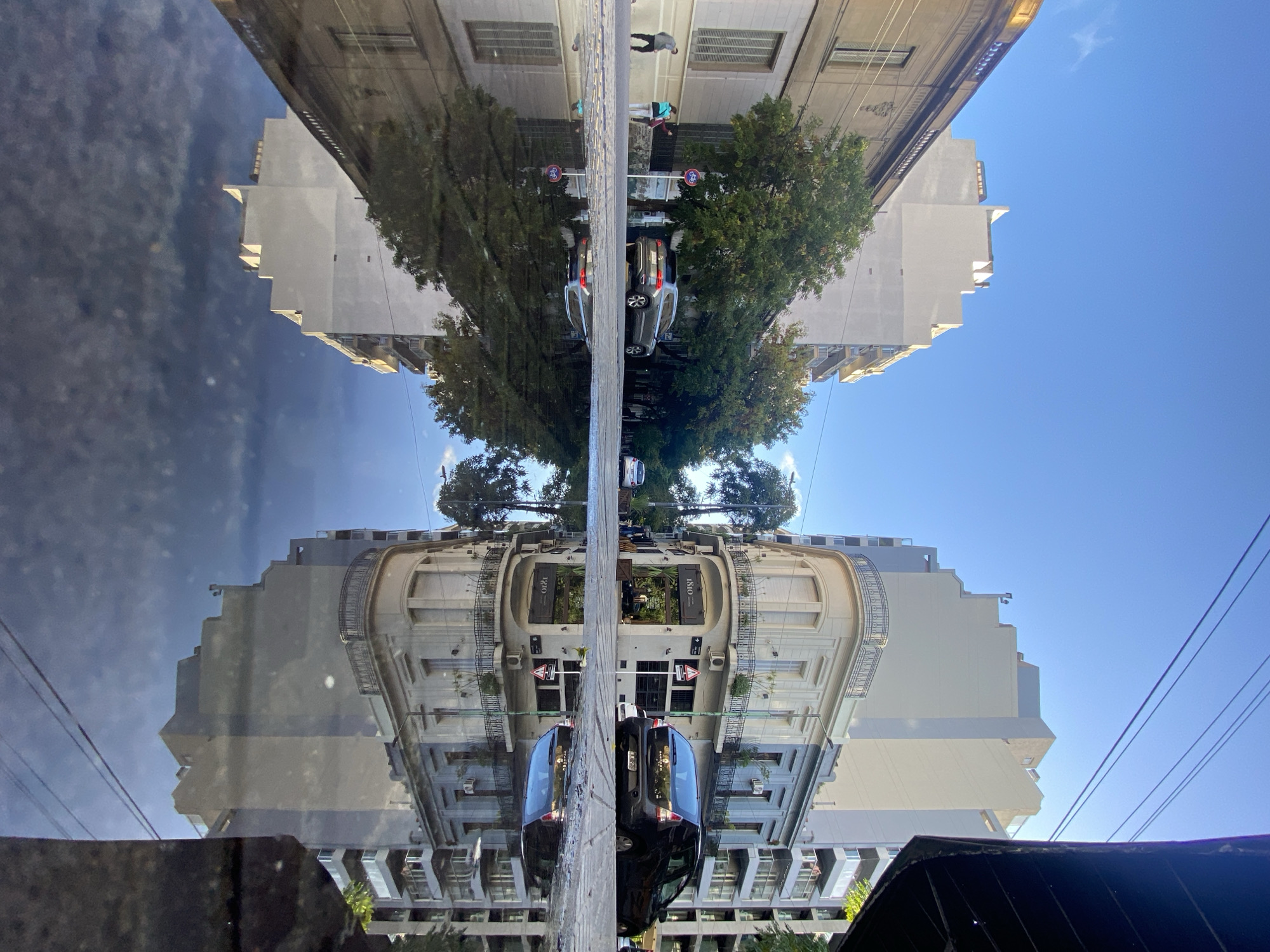 The image displays a highly symmetrical urban street scene, created by a clear reflection on a horizontal surface, likely a wet street or large puddle. The bottom half shows the actual street, while the top half presents its inverted mirror image.
The scene features a street lined with multi-story buildings on both sides. On the right, a building with ornate balconies and architectural details is prominent, displaying the year "1810" above double doors, possibly indicating a historical or themed establishment like a restaurant. Other buildings range from classic to more modern designs. Green-leafed trees are planted along the sidewalks, adding natural elements to the urban setting.
Several vehicles are visible; a dark-colored car is parked on the right side of the street in the foreground, and a silver-grey car is further down. Pedestrians are present on the sidewalks; one person in dark pants and a light top walks away on the right, and another is visible in the distance on the left. Small circular signs, including one with a bicycle symbol, are also seen. Utility wires crisscross against the sky.
The time of day is daytime, with bright sunlight and a largely clear blue sky, though some scattered clouds are reflected on the top left. The reflective surface of the street, acting like a mirror, captures all details—buildings, trees, cars, people, and sky—with remarkable clarity. Given the context, the architecture and the "1810" text strongly suggest the location is Buenos Aires, Argentina.