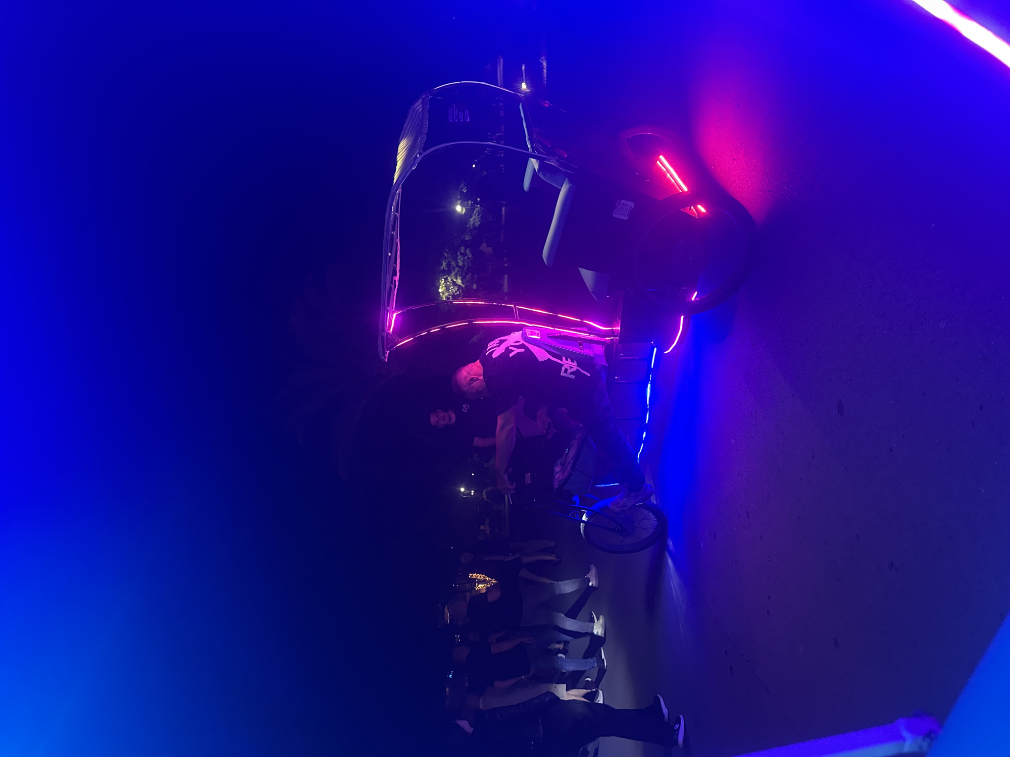 An illuminated, three-wheeled electric vehicle is parked outdoors at night, possibly in the city of Varna, Bulgaria, given the context. The vehicle features pink neon lights around its canopy and undercarriage, and red lights on its rear. A man in a black t-shirt with "REVOLT" printed in white on the back is seated in the driver's position. Several other individuals, dressed casually, are standing and walking in the background, illuminated by ambient lighting and the vehicle's lights. The ground appears to be asphalt or pavement, and there are some trees and streetlights visible in the distance. The overall atmosphere is dark with vibrant, artificial lighting, suggesting a nighttime event or gathering.