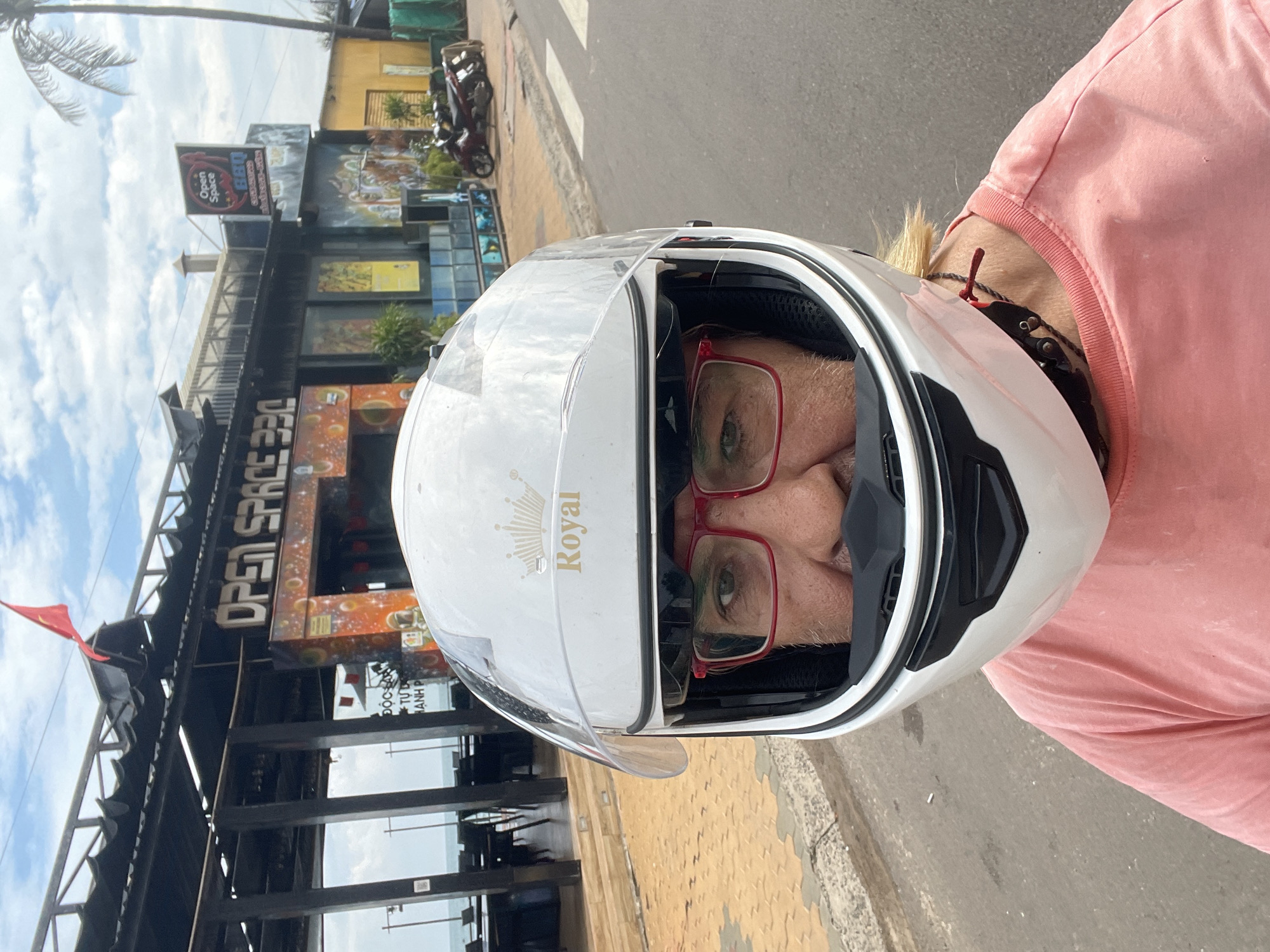 This image, presented as if correctly oriented, shows a person wearing a white motorcycle helmet and a pink t-shirt, looking directly at the viewer. The helmet features "Royal" written in gold script with a crown logo on its side. Red-rimmed glasses are visible through the helmet's clear visor, framing the person's neutral eyes. Blonde hair is visible at the back of the neck, along with a dark necklace that has a small red adornment.
The background depicts a vibrant street scene during what appears to be daytime under a partly cloudy sky. To the left, several multi-story buildings line the street, suggesting a commercial area in Phu Thuy Ward, Vietnam. A prominent sign on one building reads "Open Space" and "B&Q", with additional Vietnamese text beneath, which includes phrases resembling "độc đáo" (unique) and "sáng tạo" (creative). A lower sign clearly displays "OPEN SPACE 330". A red flag, possibly the national flag of Vietnam, flies from one of the structures. Palm tree fronds are visible against the sky on the far left. The scene includes a paved road and a yellowish-tiled sidewalk. Parked motorcycles are visible further down the street, indicating common local transportation.