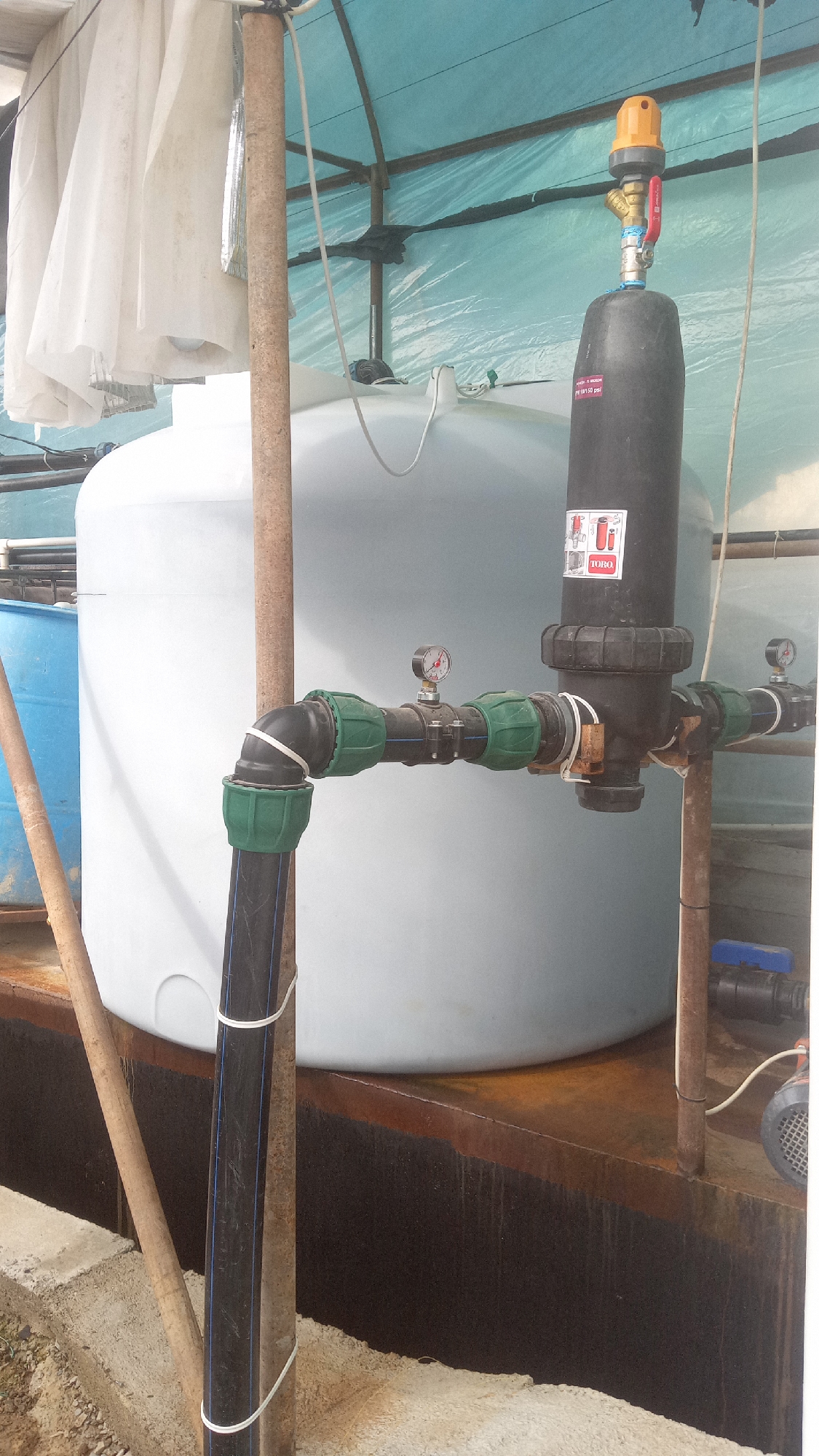 This image depicts an industrial water filtration system located in Salar, Uzbekistan. The system features a large white plastic water tank, a black filter housing with pressure gauges, and various pipes and connectors. A blue barrel is partially visible to the left of the white tank. The setup is situated under a light blue tarp structure supported by metal poles. Wooden stakes and concrete structures are also visible in the foreground, suggesting an outdoor or semi-outdoor industrial setting. The lighting indicates it is daytime. There are no people visible in the image.
