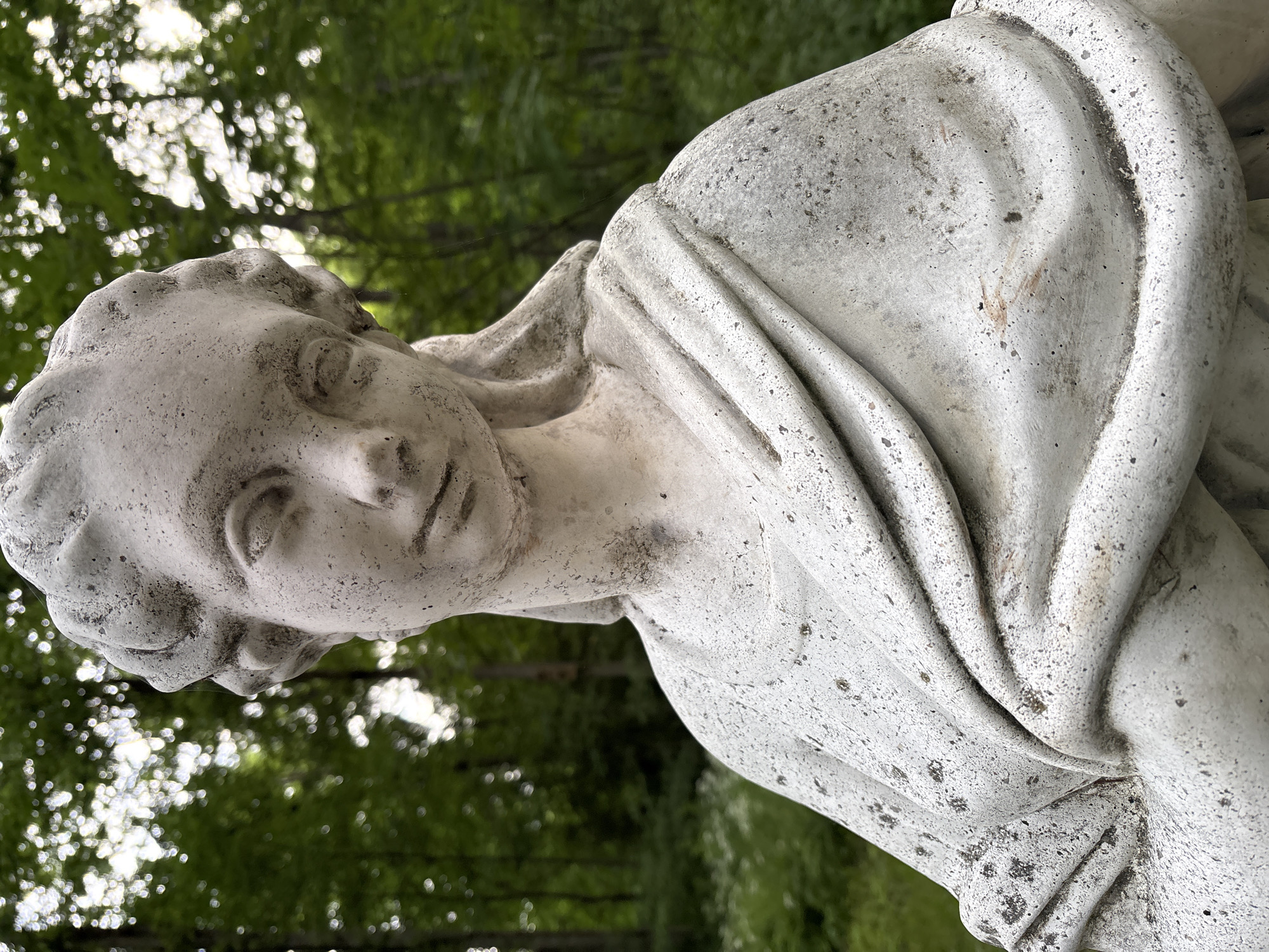 The image is a close-up, eye-level shot of a weathered, white stone statue of a young woman. The statue is outdoors, with a blurred background of lush green trees and dappled sunlight filtering through the leaves. The statue's head is slightly tilted down and to the left, with a serene or pensive expression. Her hair is styled in curls around her head, and she wears a draped garment that covers her shoulders and chest. The stone is textured and shows signs of age, with some dark staining and pitting. The statue is positioned in what appears to be a garden or wooded area, with hints of white flowers or foliage in the lower left background. The specific location within the city of Gorki, Russia, is not provided in the image.