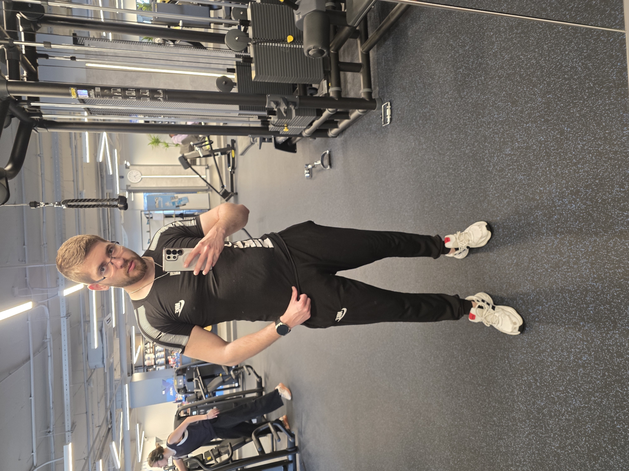 A man is captured in a mirror selfie inside a modern gym, looking directly at the camera. He stands upright, holding a light-colored smartphone to take the picture. He has short brown hair, a beard, and wears glasses. His attire consists of a black t-shirt with white stripe details on the short sleeves and a white "NIKE" logo on the chest, paired with black sweatpants that also feature a white Nike logo on the left thigh. On his left wrist, he wears a black smartwatch. His feet are clad in white and red sneakers.
The background, reflected in the mirror, reveals a well-lit gym environment with bright linear light fixtures on the ceiling. Various pieces of exercise equipment are visible, including a large cable machine with weight stacks. A smaller dumbbell lies on the dark grey gym floor, which is speckled with lighter blue and white dots. In the reflection, another person, likely a woman, is seen in the mid-ground, focused on exercising on a leg machine. She is wearing dark activewear and appears to be wearing headphones. The gym appears spacious and well-maintained. The man's expression is neutral, and the overall scene depicts a typical gym setting during an exercise session.