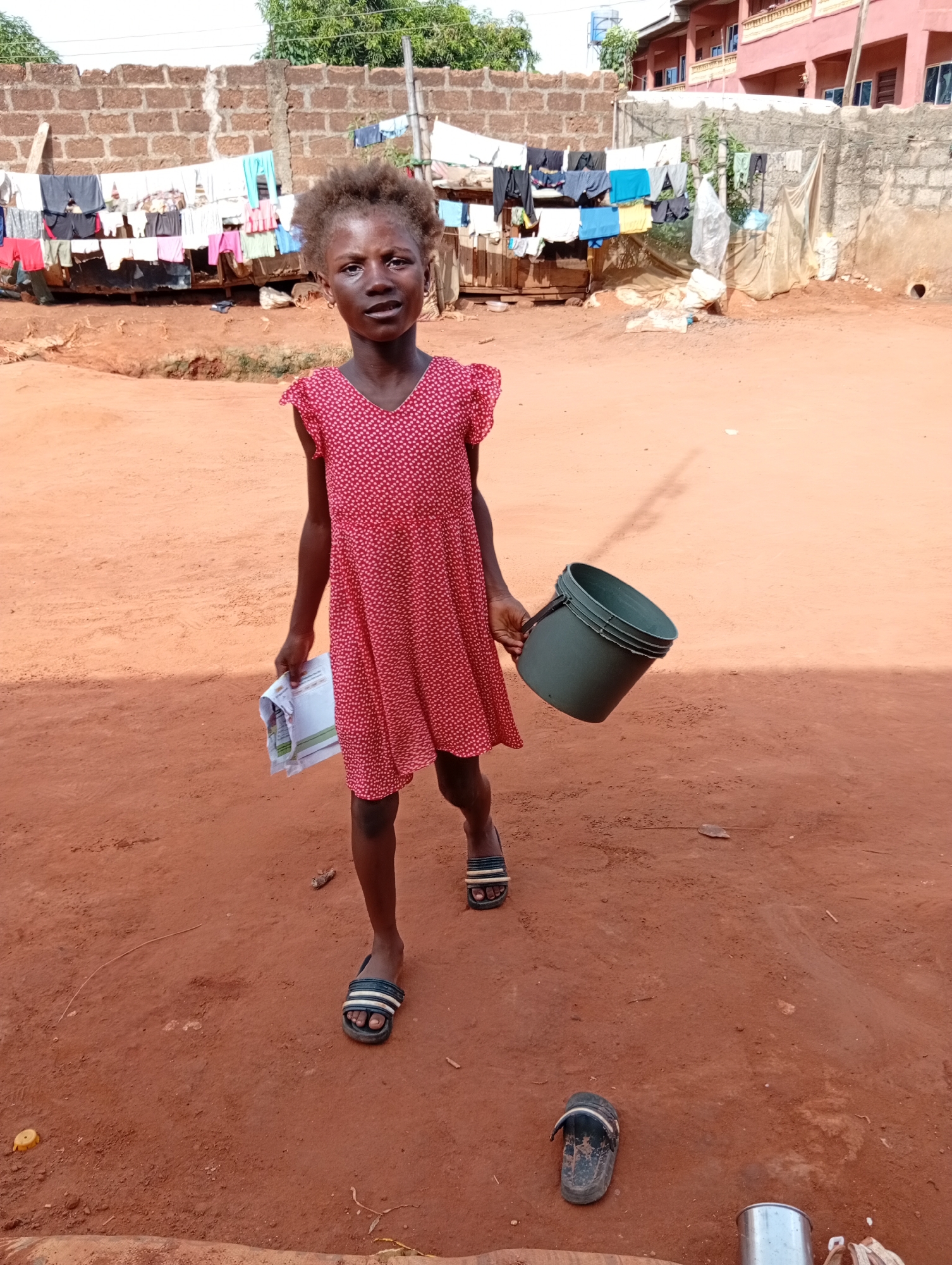 A young girl stands centered in an outdoor, unpaved area in Ogidi, Nigeria. She has dark skin and kinky dark hair. She is wearing a red, short-sleeved dress with small white polka dots and dark-colored slide sandals with white stripes; one sandal is on her left foot, while the other is on the reddish-brown dirt ground near her right foot. In her left hand, she holds a stack of white papers or booklets that appear to have some text, and in her right hand, she carries a dark green plastic bucket. She looks directly at the viewer with a neutral, slightly solemn expression.
The scene is brightly lit by daylight, indicating a sunny day. The ground is composed of reddish-brown earth. In the background, a low wall made of reddish-brown concrete blocks spans the mid-ground. Behind this wall, several clotheslines are visible, laden with numerous items of laundry—shirts, trousers, and other garments in various colors—hanging to dry. Further back, on the right side of the frame, a multi-story residential building with a reddish-pink exterior, featuring multiple windows and balconies, stands prominently. Another wall, constructed from grey concrete blocks, is situated in front of this building. Green foliage is visible above the walls. A silver metal container or cup is on the ground to the lower right. The overall setting suggests a community environment.