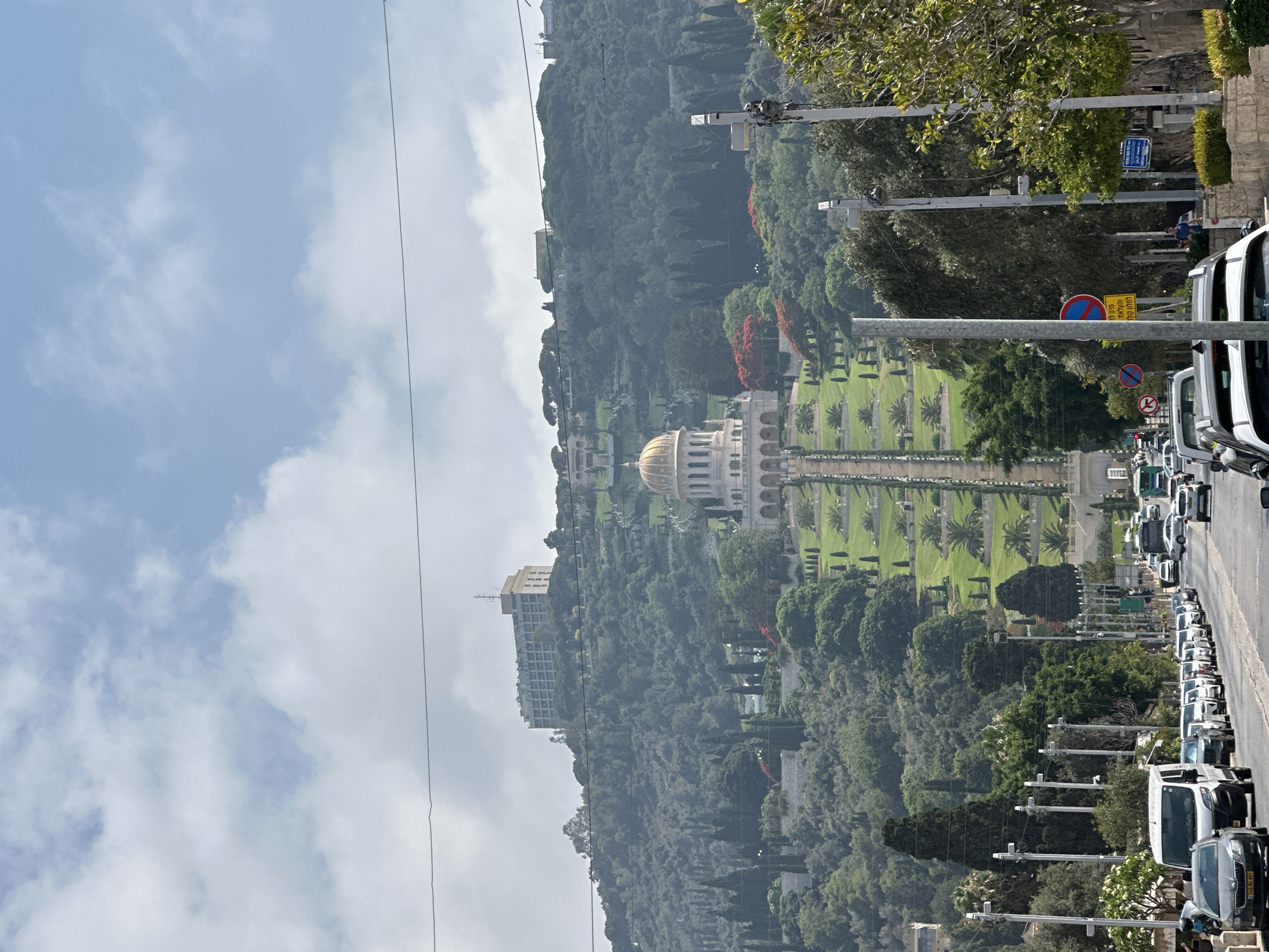The image captures a view of the Baháʼí World Centre in Haifa, Israel, with a street in the foreground. The Baháʼí structure, characterized by its golden dome, is prominently situated on the lush green hillside. The hillside is terraced with meticulously maintained gardens and pathways leading up to the building. A large hotel-like structure is visible further up the hill. The street in the foreground is lined with parked cars and trees, with a few vehicles in motion. The sky above is partly cloudy. The scene appears to be captured on a clear day, with good visibility.