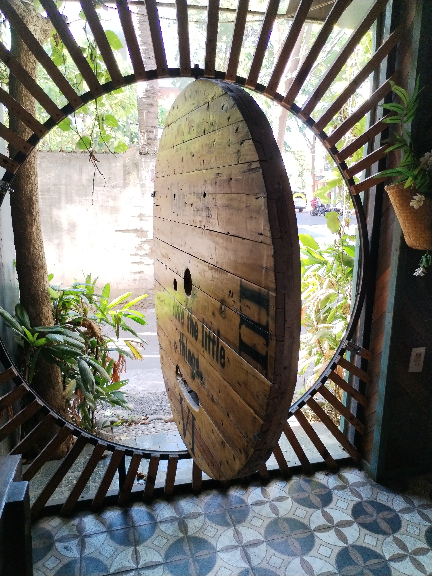 The image shows an indoor setting in Bac Nha Trang Ward, Vietnam. A large, wooden cable spool dominates the view, partially obstructing the outside scene. The spool has several holes and lettering that reads "love the little things". The spool is positioned within a circular, lattice-like structure made of wood. The exterior visible through the gaps in the structure shows greenery, a street, and a motorbike with two people on it. The foreground features patterned tiles in blue and gray. To the left, a tree trunk and foliage are visible. To the right, a wall with a hanging basket containing plants is present.