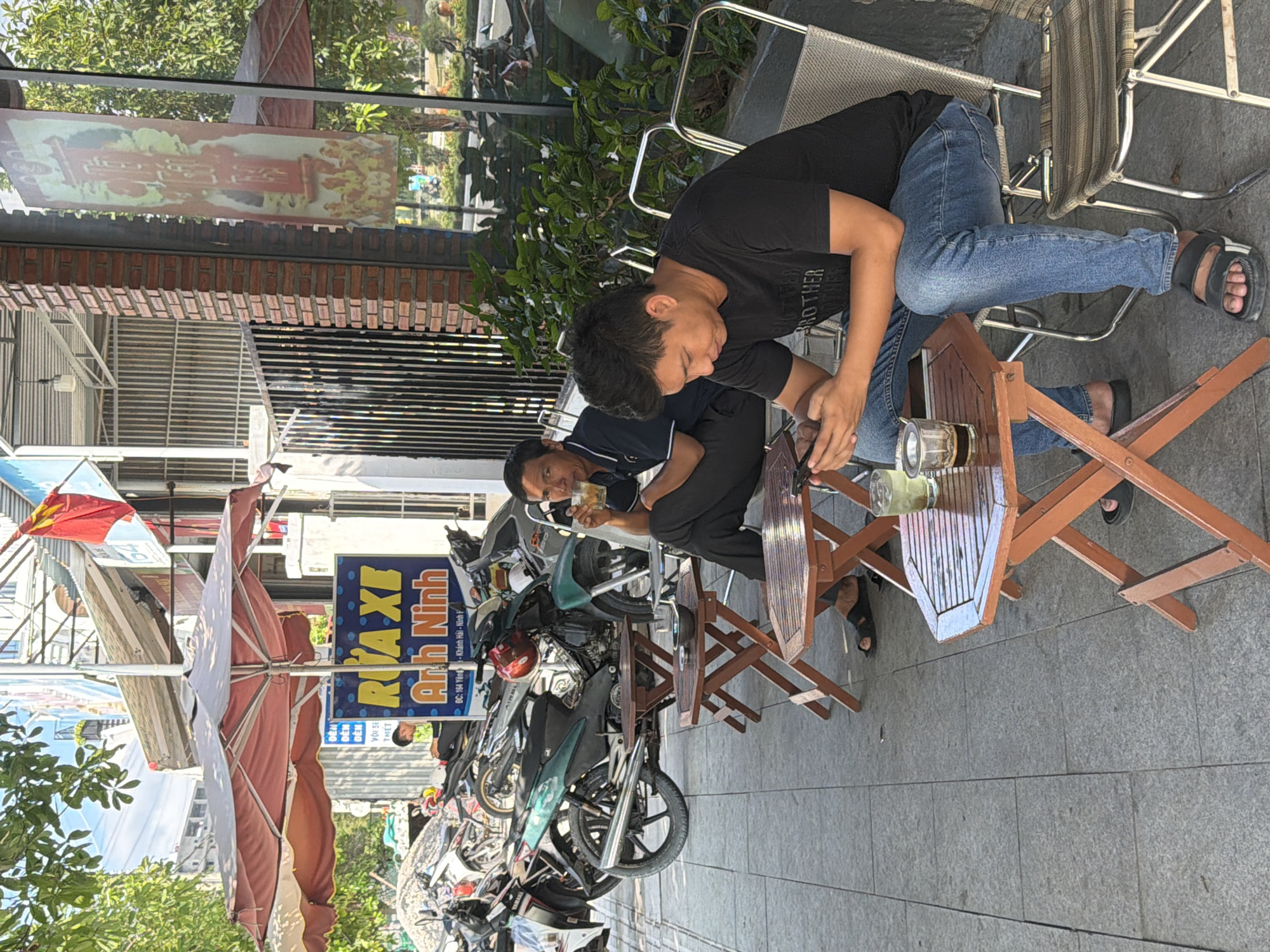 This image captures a vibrant street scene in Ninh Chu Ward, Vietnam, depicting two men relaxing at an outdoor cafe. The setting is a bustling sidewalk lined with parked motorcycles and small, wooden tables and chairs.
In the foreground, a man wearing a black t-shirt with the word "BROTHER" visible, and blue jeans, is seated at a small slatted wooden table. He appears to be looking down at something in his hands, possibly a phone. A glass with a dark beverage is on his table. Slightly behind him, another man in a dark polo shirt and dark pants is also seated at a similar table, holding a glass containing a lighter, possibly green, liquid. The overall atmosphere suggests a casual break or social interaction.
Several motorcycles are parked along the curb, a common sight in Vietnamese street environments. A large, reddish-brown parasol is partially folded, providing shade over the outdoor seating area. The background features buildings with brick facades and some lush green foliage. Bright daylight and clear shadows indicate it is daytime, likely mid-morning or afternoon.
Visible text in the image includes "BROTHER" on the t-shirt of the man in the foreground. A blue sign reads "RUA XE Anh Ninh" which translates to "Motorbike Wash Anh Ninh," followed by an address: "ĐC: 164 Yên - Khánh Hải - Ninh H", confirming a local Vietnamese business. Other less clear text like "DEN" and "THET" also appears on other signs. The scene portrays a typical moment of daily life in a Vietnamese urban area.