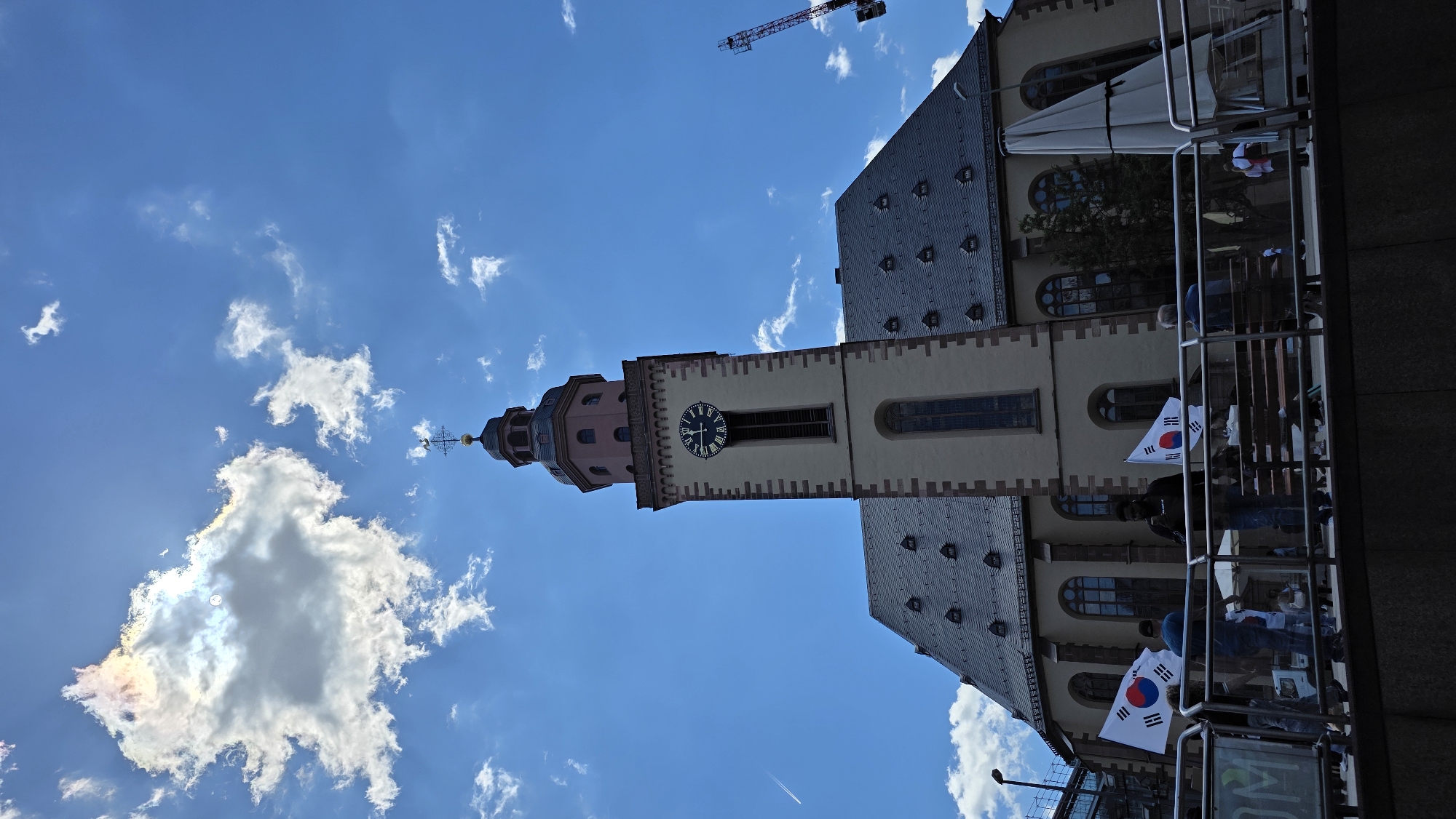 In an outdoor urban setting, a large church building with a prominent clock tower dominates the scene. The sky above is a bright blue, punctuated by scattered white clouds, one of which exhibits iridescent colors. A contrail from an airplane is visible in the upper left quadrant of the sky. The church features a dark gray tiled roof with numerous dormer windows and a tall, pale yellow clock tower with a dark clock face. At the base of the church, in the foreground, a metal railing separates the viewer from a group of people. Several individuals are gathered, with some holding up flags of South Korea. The flags are white with a red and blue yin-yang symbol and black trigrams in the corners. The people appear to be standing on a raised platform or walkway. In the background, a construction crane is visible. The overall impression is of a sunny day in a European city, with a public gathering or event taking place in front of a historic church.