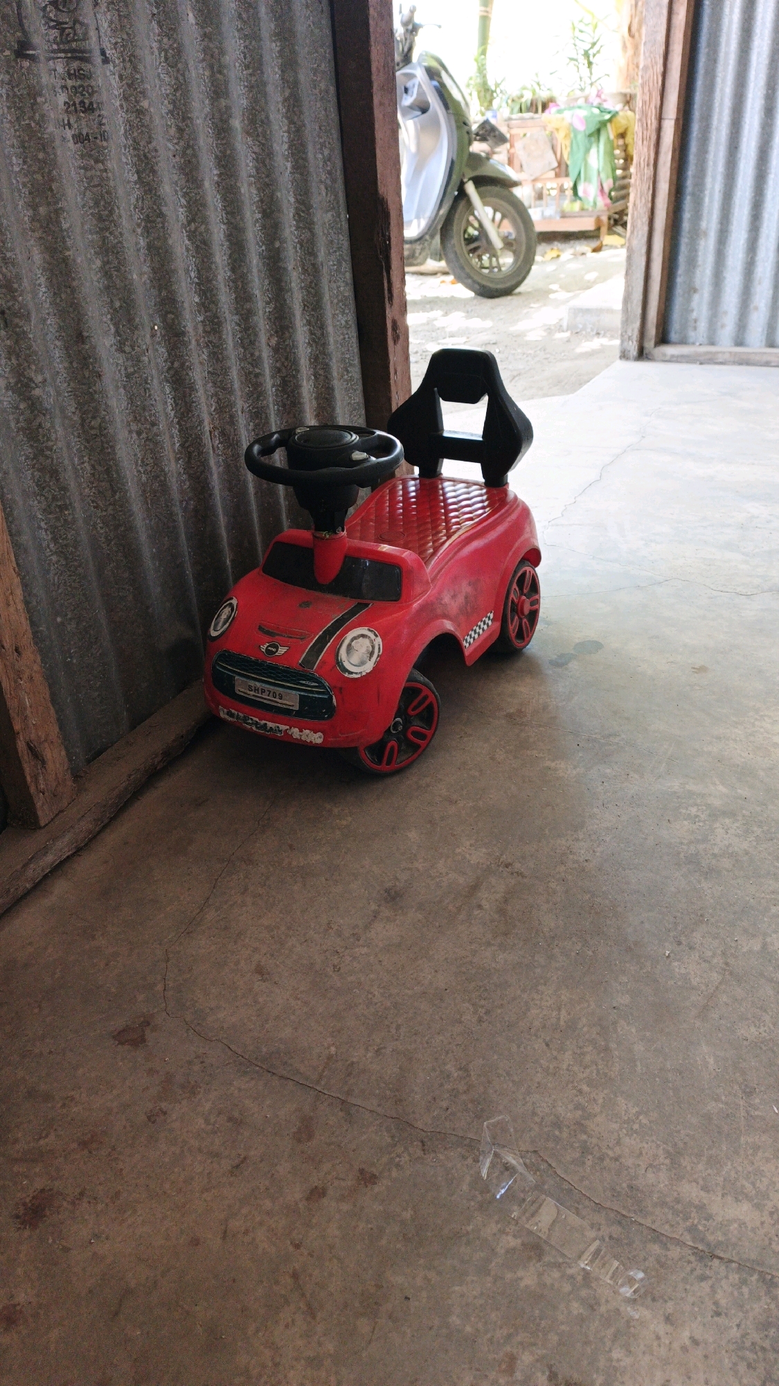 The image depicts a red toy car parked on a concrete floor next to a corrugated metal wall. A scooter is visible in the background, partially obscured by an open doorway. The scene appears to be outdoors or in an open-air structure. The toy car is a miniature replica of a car, complete with a steering wheel and wheels. The corrugated wall is weathered and appears to be part of a shed or workshop. The concrete floor is cracked in places, and there are some dark stains on it. A transparent plastic object, possibly a broken piece of packaging or a cup, lies on the floor in the foreground. The lighting suggests it is daytime. This image does not provide specific information about the city of Kiping, Indonesia, beyond what can be inferred from general environmental cues.