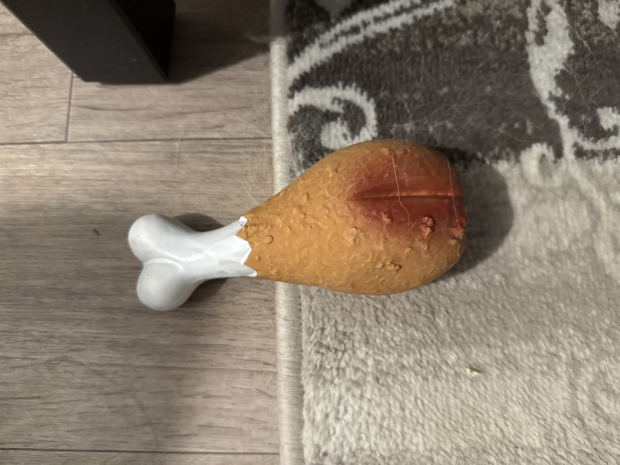 A dog toy shaped like a chicken leg rests on a wood-grain floor and a patterned rug. The toy has a white, bone-shaped end and an orange, textured body. A dark, rectangular object is partially visible in the upper right corner of the image. The scene does not provide any information about the city of Perm, Russia.