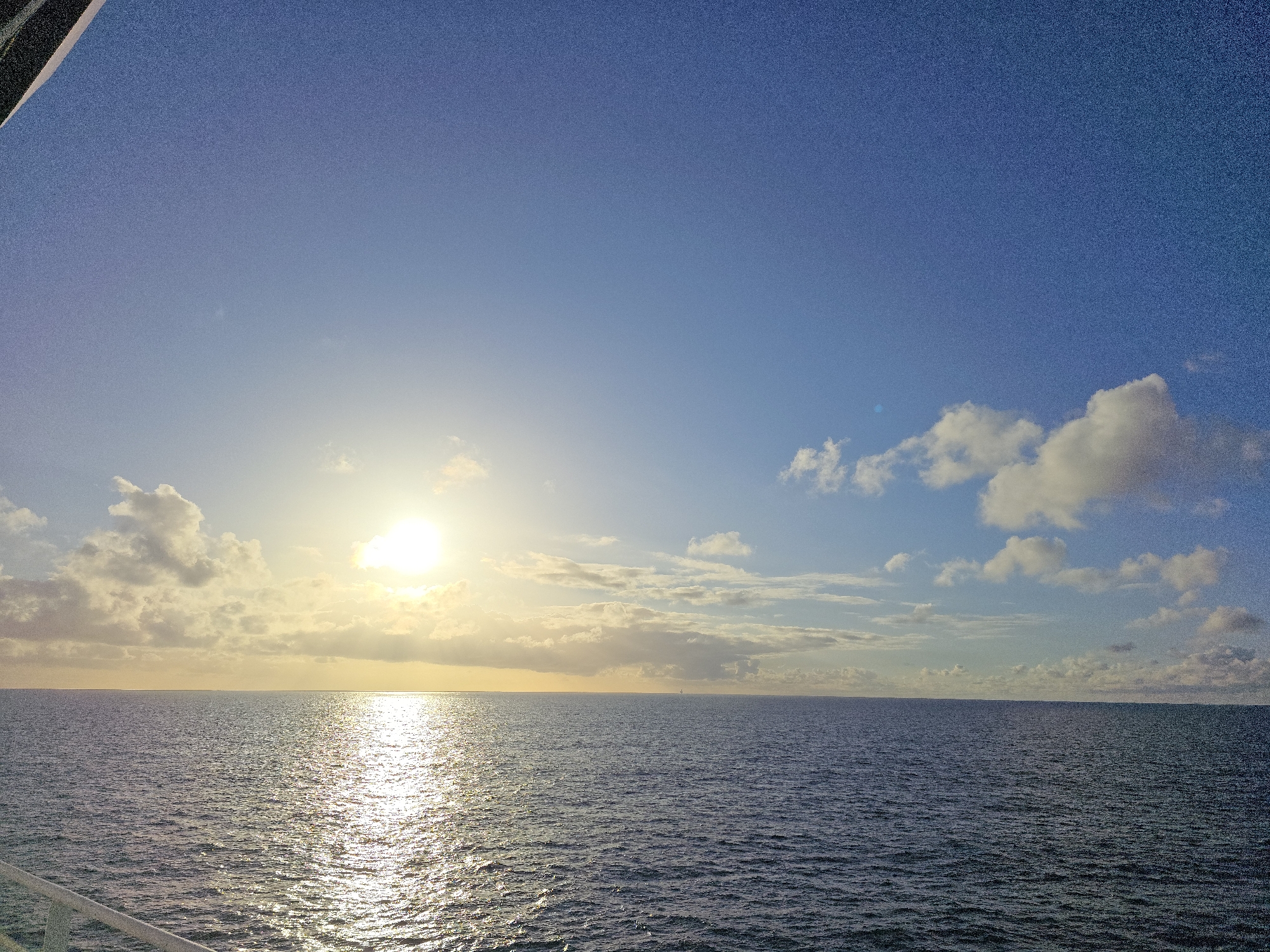 The image shows a view of the ocean under a sky with scattered clouds and the sun. The sun is positioned low in the sky, creating a bright reflection on the water's surface. The sea appears calm with gentle ripples. The sky is a mix of light blue and white, with some darker blue patches. There is a railing visible in the lower-left corner, suggesting the photo was taken from a boat or ship. The horizon line is relatively straight and even.