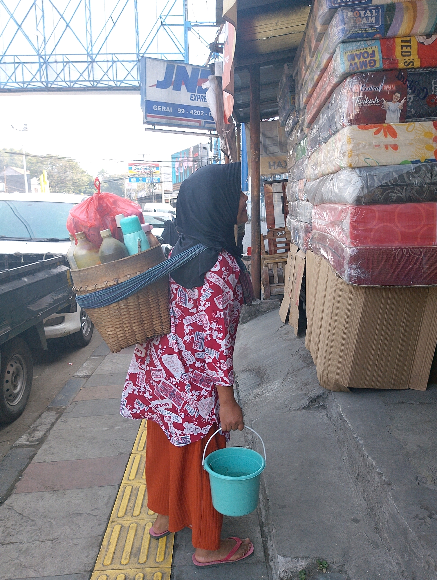 A person wearing an orange skirt and a red and white patterned blouse, and a black hijab, stands on a sidewalk in Lembang, Indonesia, holding a light blue bucket with a white handle. They are carrying a woven basket on their left shoulder containing bottles and a red plastic bag. Behind them, a stack of mattresses wrapped in plastic is visible, along with a partial view of a vehicle. A sign for "JNE EXPRESS" is seen in the background. The environment appears to be an urban street with a concrete sidewalk and road.