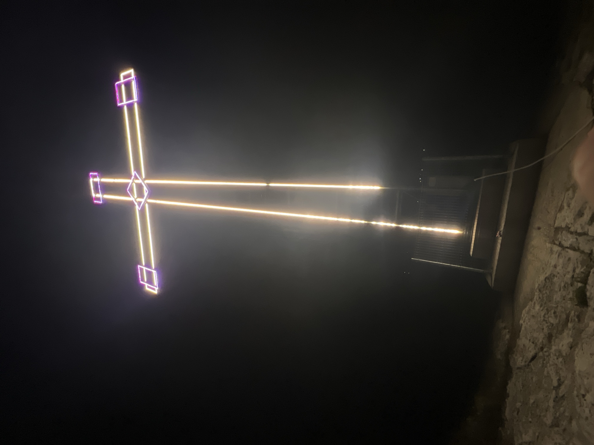 An illuminated cross stands tall against a dark, foggy sky. The cross is outlined with neon lights, predominantly yellow, with accents of purple at the ends of the horizontal beams and the top of the vertical beam. The light from the cross creates a hazy glow in the foggy atmosphere. At the base of the cross, there is a structure with steps and a metal fence. The ground in front of the structure appears to be rocky or uneven. The overall scene suggests a nighttime setting, possibly on a hill or elevated area, given the darkness and the visible structure at the base. The image is taken from a low angle, looking up at the cross. The location is identified as the city of Provadia, Bulgaria.