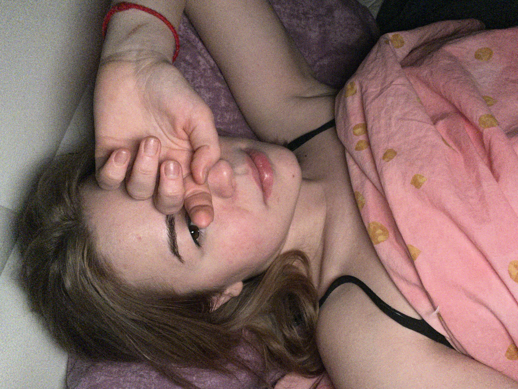 A young female subject is depicted in an indoor setting, identified as Boreti, Montenegro, lying in a bed. She is positioned with her head on a purple pillow, while her body is partially covered by a pink blanket adorned with irregular yellowish patterns. Her right hand is raised, fingers slightly splayed, partially obscuring her forehead and the area around her right eye, which is visible and directed towards the viewer. A red string bracelet is present on her left wrist. She is wearing a black strap garment. The image exhibits a noticeable grain, consistent with low light conditions.