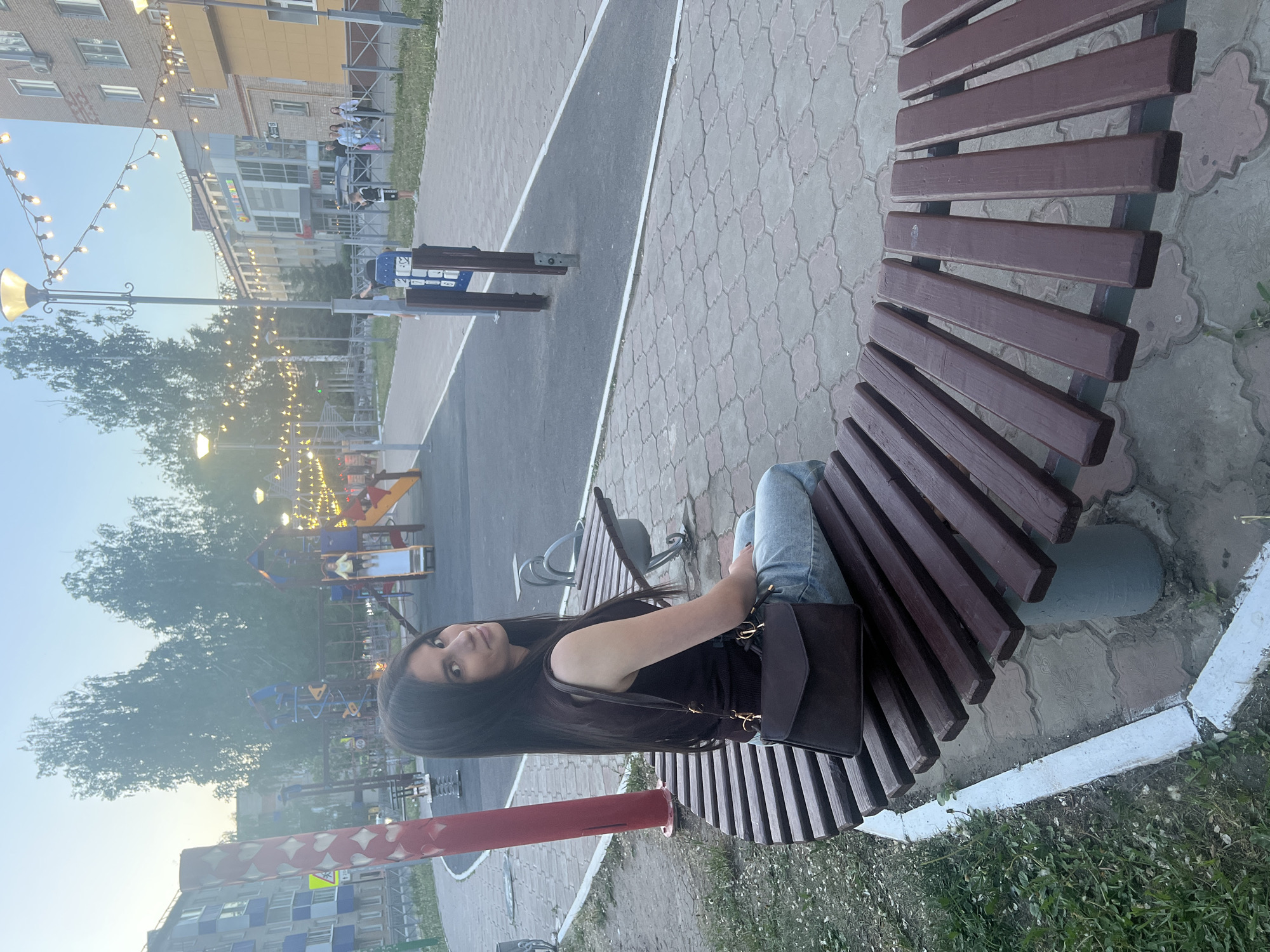 A young woman with long, dark hair sits on a curved wooden bench in a park in Mendeleyevsk, Russia. She wears jeans and a black top, with a black purse resting on the bench beside her. In the background, a playground is visible, illuminated by string lights. There is a pathway and buildings in the background.