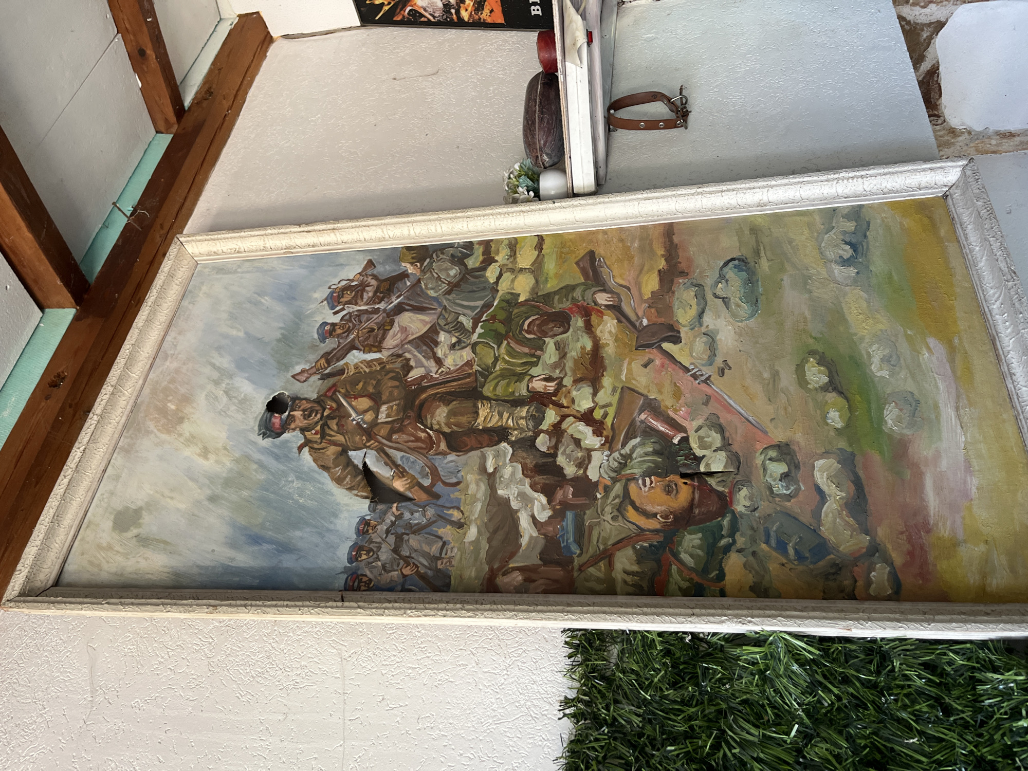 A framed painting of soldiers in battle hangs on a white wall. The painting depicts soldiers in various shades of brown, green, and blue amidst a landscape of greens, browns, and yellows. The lower part of the image is partially obscured by green, possibly artificial, foliage. Above the painting are ceiling beams and other wall-mounted objects. The location is Unknown, Unknown.