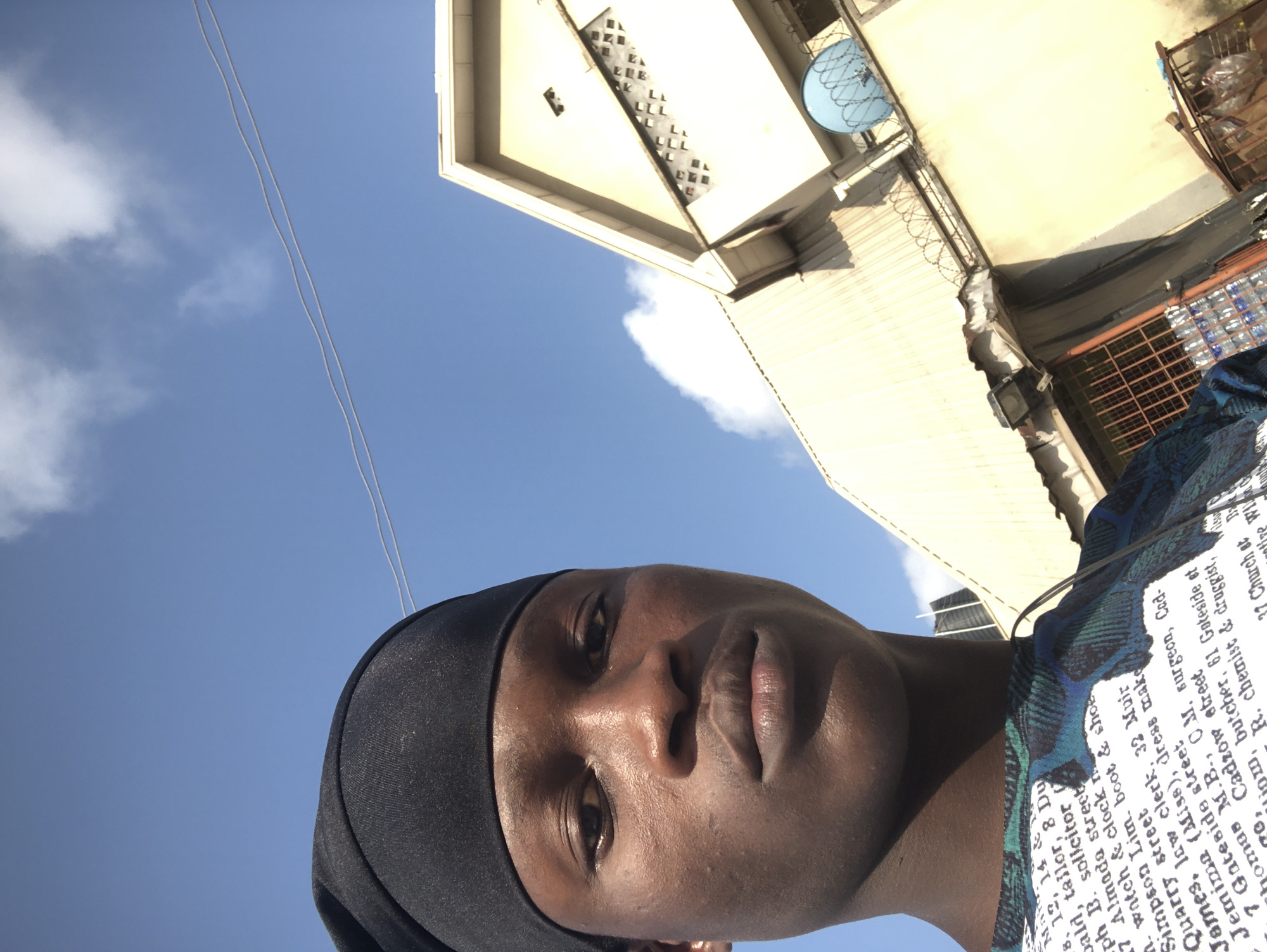 A young Black male wearing a black head covering and a patterned shirt poses for a selfie outdoors, looking directly at the camera. The background features a light-colored building with a blue satellite dish and a clear blue sky with scattered clouds. Text is visible on the man's shirt, appearing to be a list of names and addresses, though the exact wording is illegible due to resolution. The image seems to have been taken during the day, with natural light illuminating the scene. The location is identified as Ado, Nigeria.