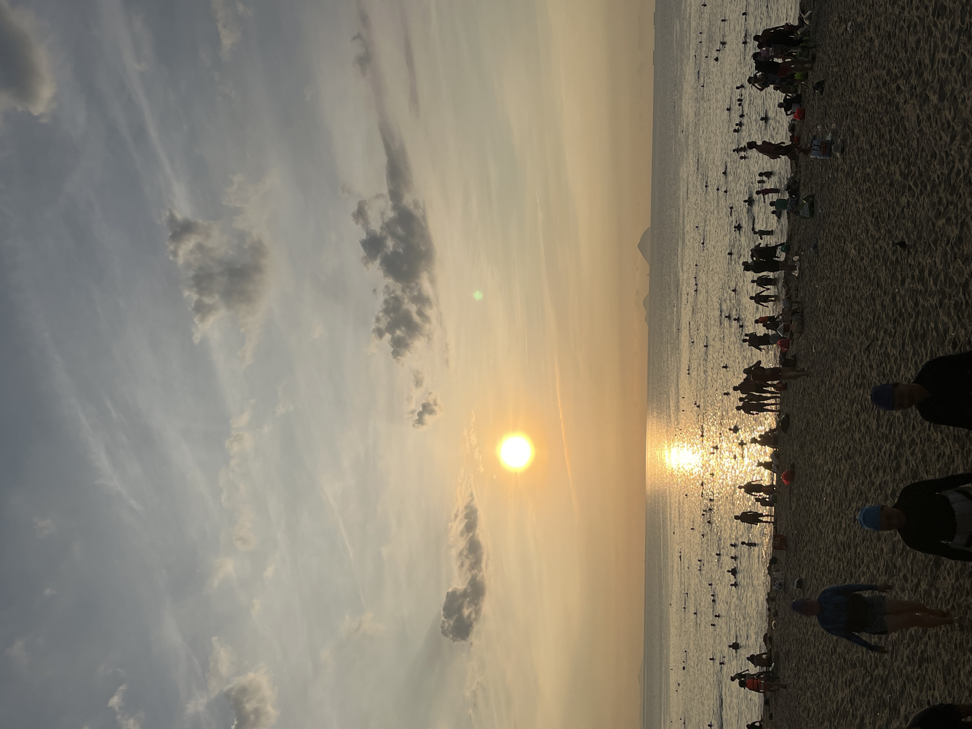 The image depicts a beach at sunset, likely in Nha Trang Ward, Vietnam. The sun is setting over the water, casting a golden glow on the scene. Numerous people are on the beach and in the water, some wearing swimming caps and appearing to be engaged in swimming or water activities. The beach is sandy and crowded, and the sky is filled with clouds. A land feature is visible in the distance.